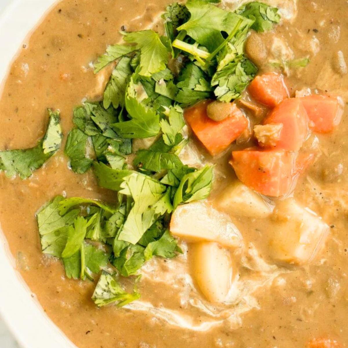 A bowl of hearty lentil soup with carrots, potatoes, and a swirl of cream. The soup is garnished with fresh cilantro.