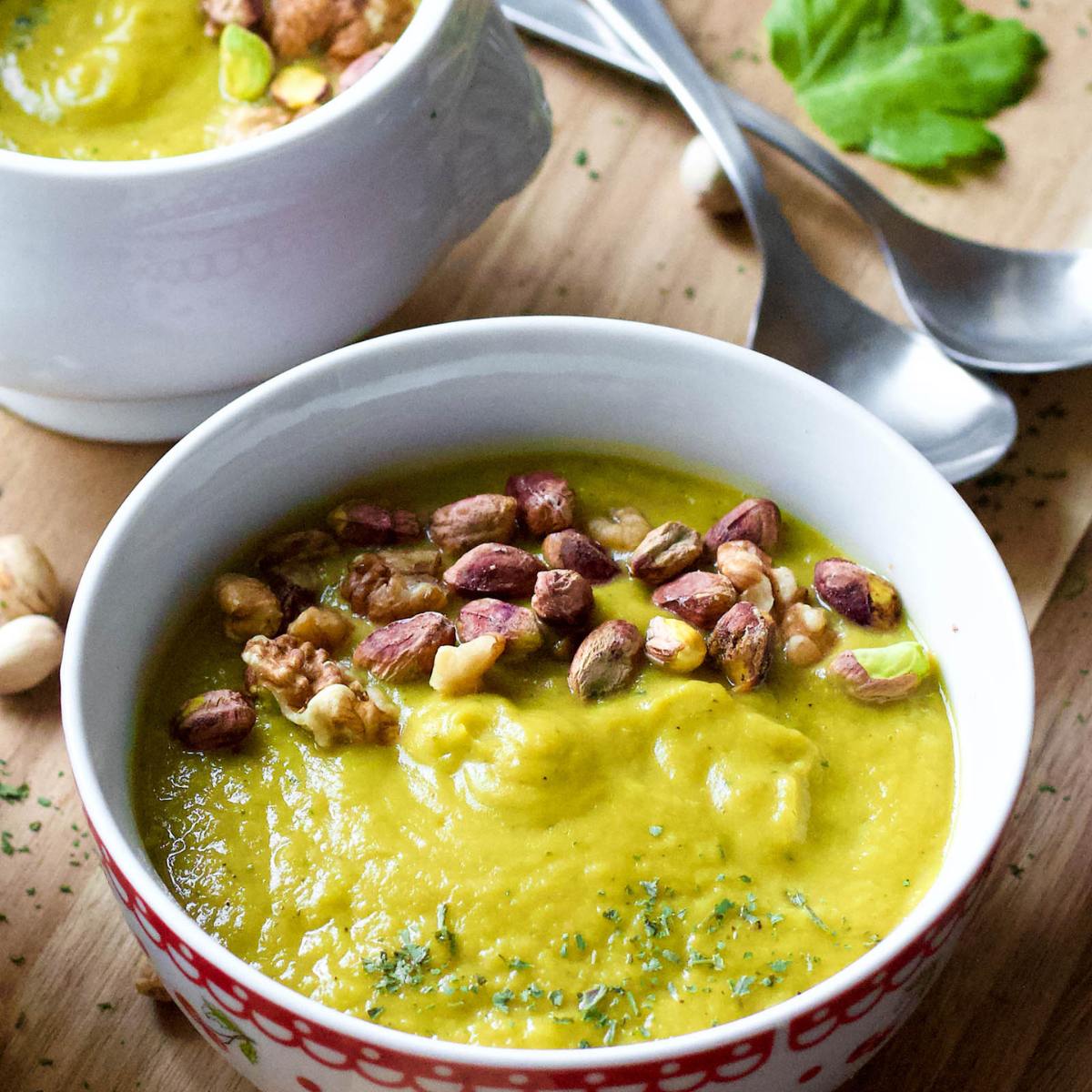 A bowl of creamy, green broccoli soup topped with chopped walnuts and pistachios. A second bowl of soup is visible in the background, along with two spoons and some scattered nuts.