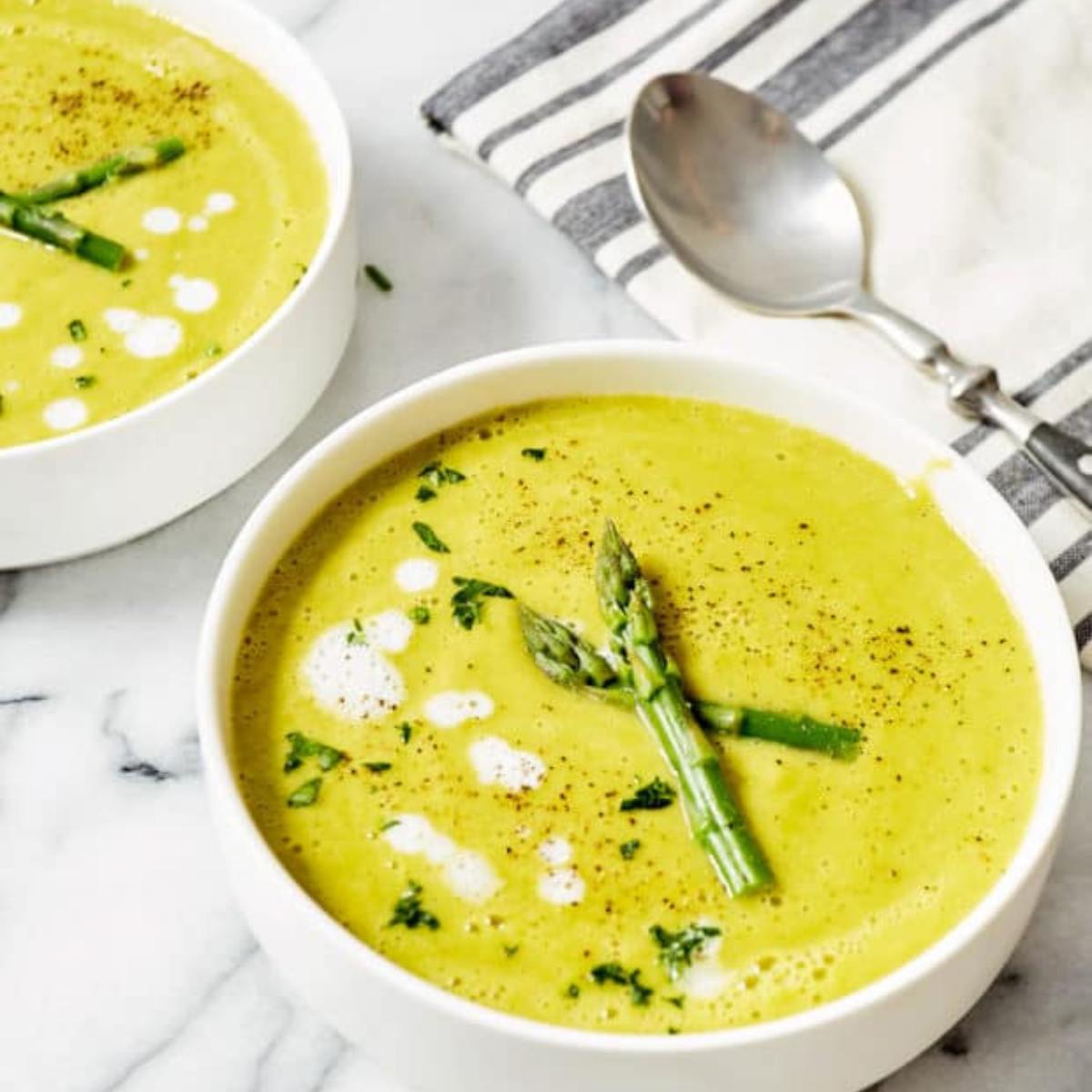 Two bowls of creamy, pale green asparagus soup. The soup is garnished with fresh asparagus spears, parsley, and a swirl of cream. A spoon is resting on a striped napkin next to the bowls.