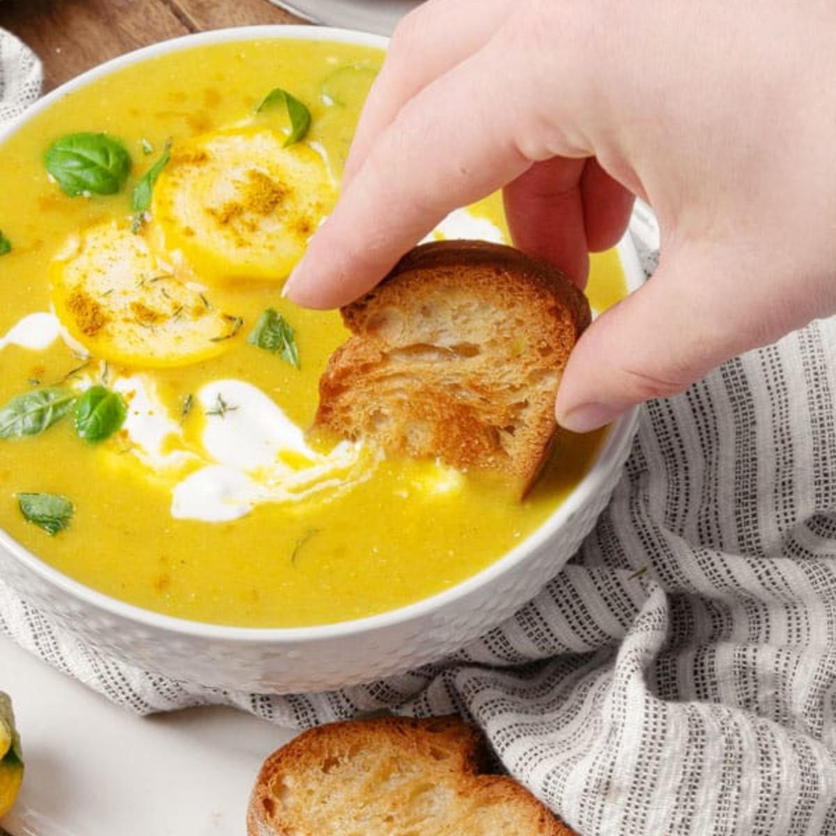 A hand dipping a piece of crusty bread into a bowl of creamy, yellow soup.