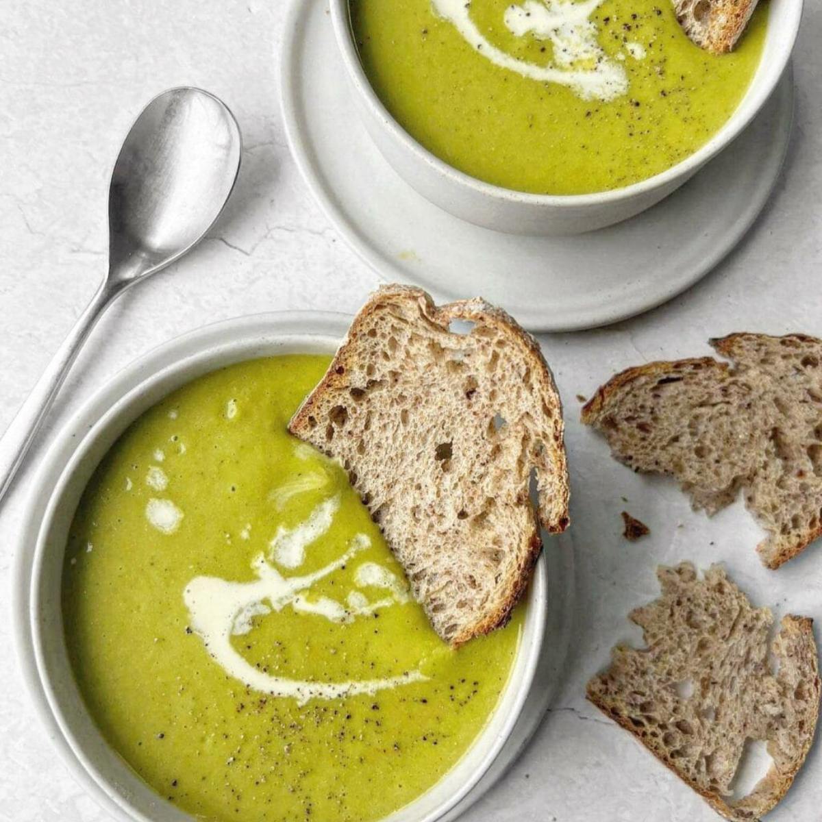 Two bowls of creamy, vibrant green soup. The soup is topped with a swirl of cream and a sprinkle of black pepper. One bowl has a slice of crusty bread resting on the rim.