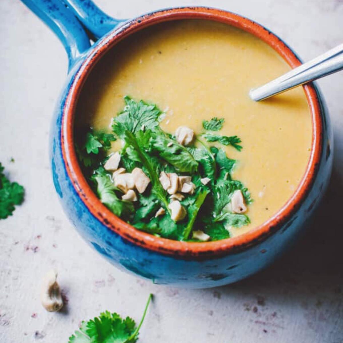 A bowl of creamy, golden soup topped with fresh cilantro and chopped cashews. The bowl is a vibrant blue with an orange rim.