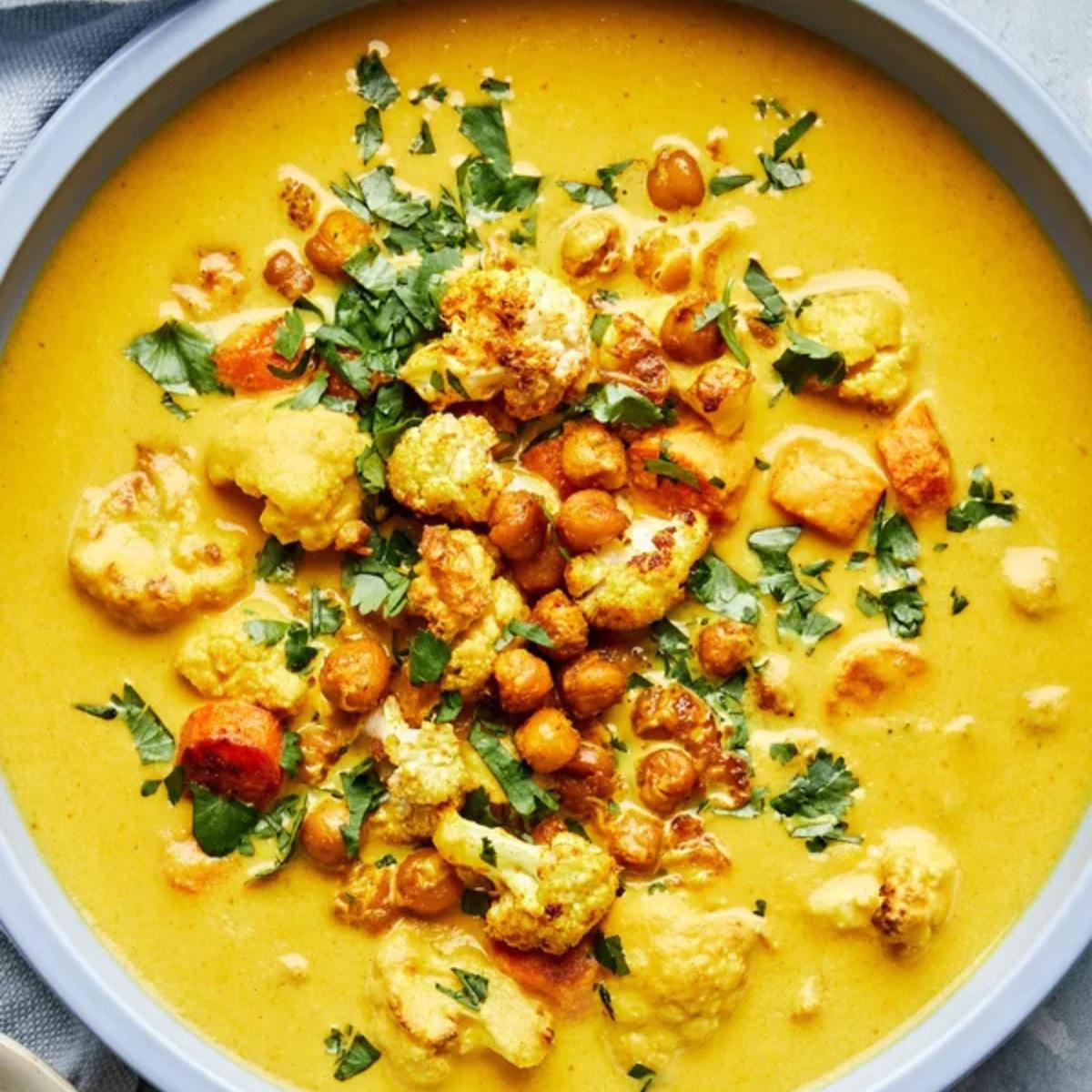 A bowl of creamy, golden curry soup with chickpeas, cauliflower florets, and carrots. The soup is garnished with fresh parsley.