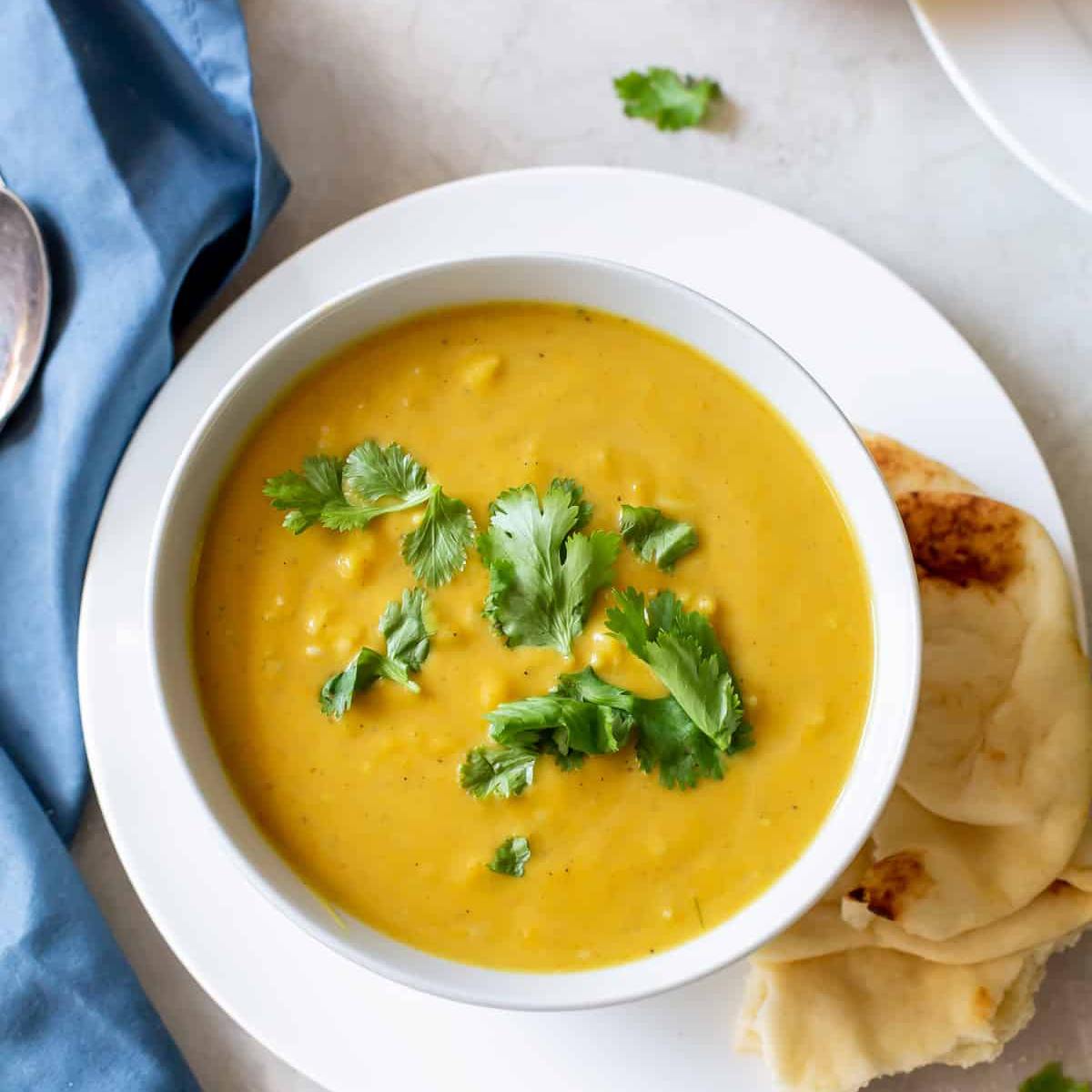 A bowl of creamy, golden lentil soup garnished with fresh cilantro. A piece of naan bread is resting on a plate next to the bowl.