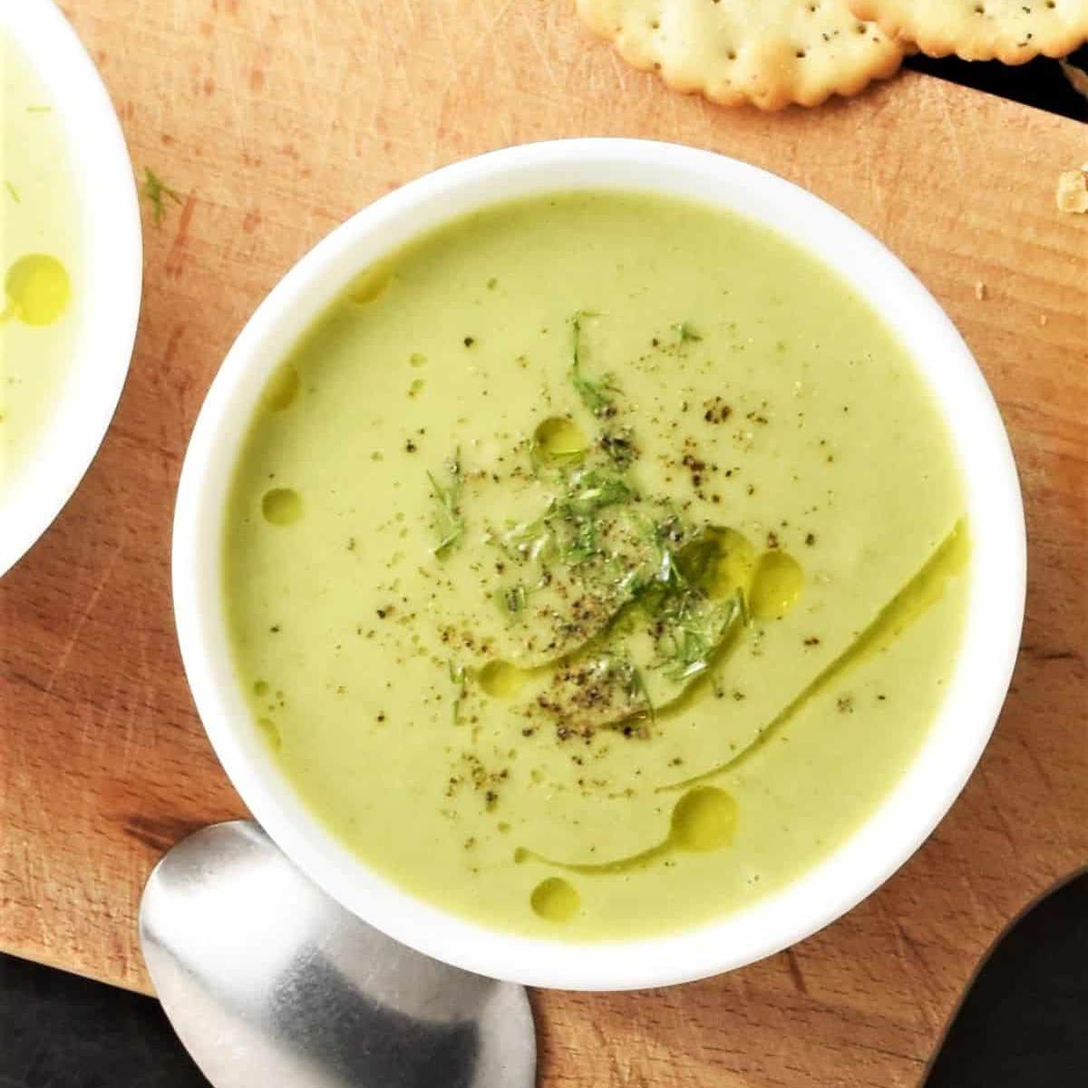 A bowl of creamy, pale green soup garnished with a drizzle of olive oil, black pepper, and fresh herbs. 