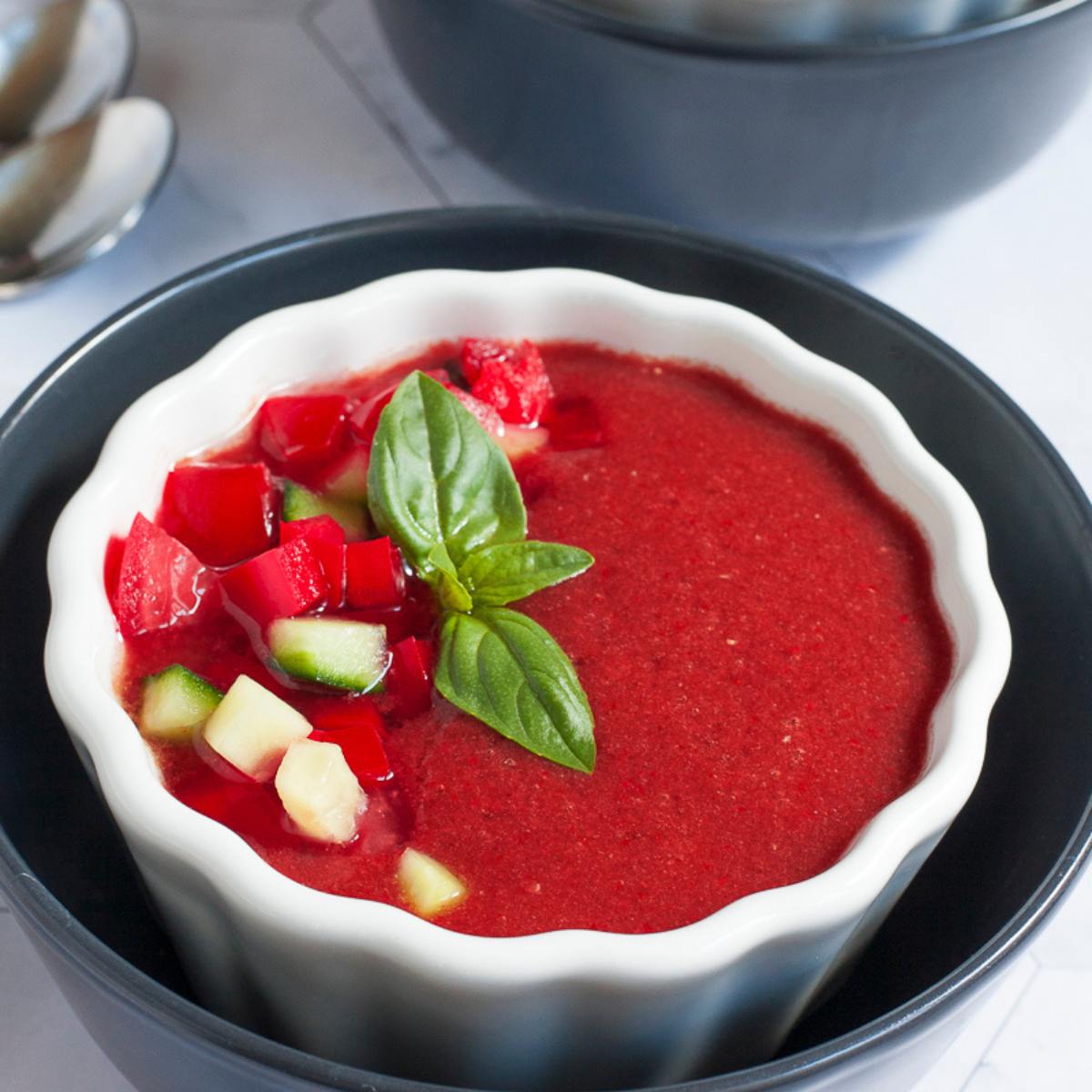 The soup is garnished with diced cucumber, chopped red pepper, and fresh basil leaves. The bowl is white with a fluted rim and is resting on a black plate.