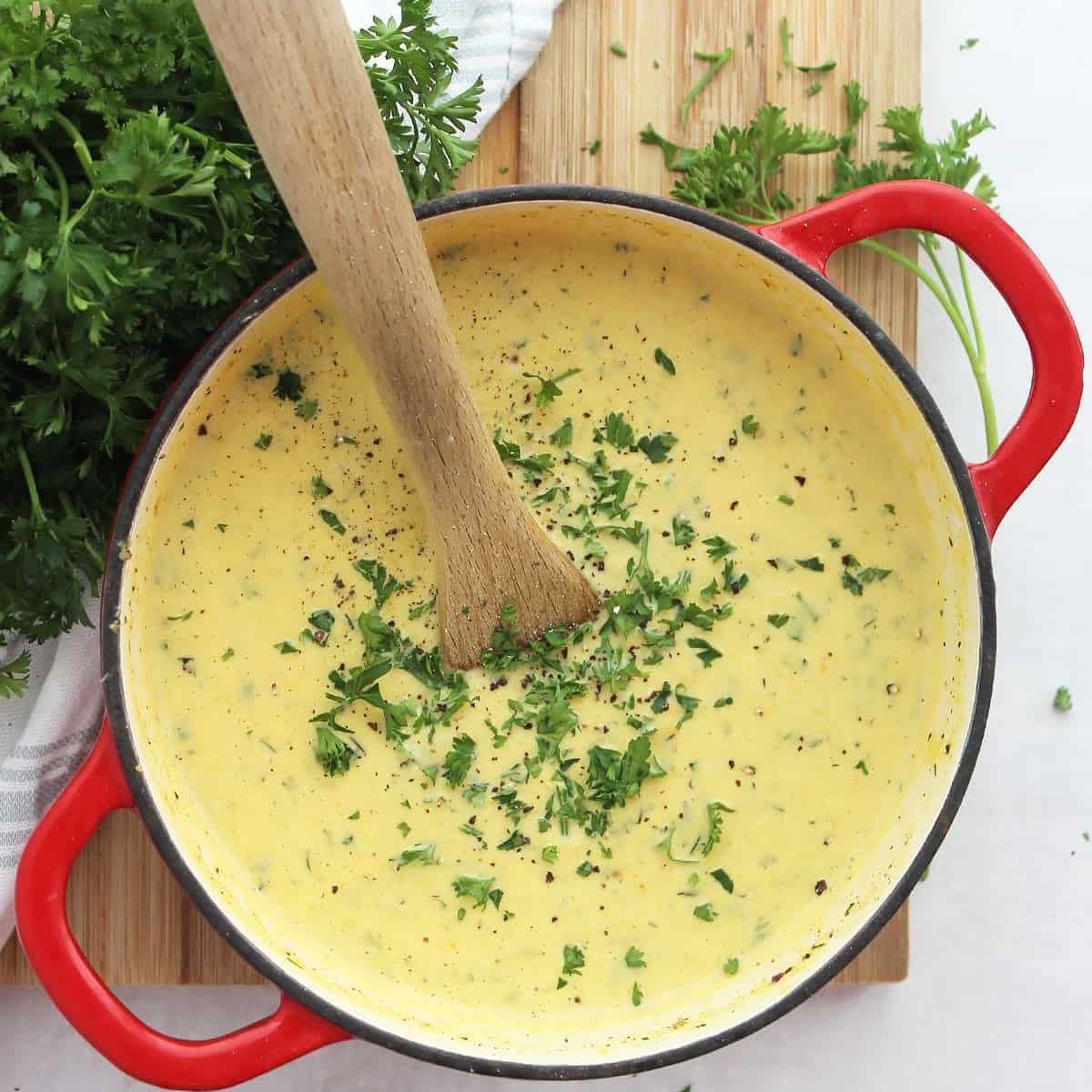 A large red pot filled with a creamy, yellow soup. Fresh parsley is sprinkled on top, and a wooden spoon rests in the pot.