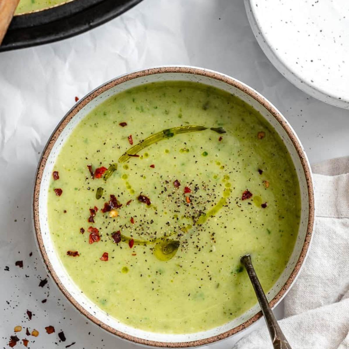 A bowl of creamy, pale green soup garnished with a drizzle of olive oil, black pepper, and red pepper flakes.