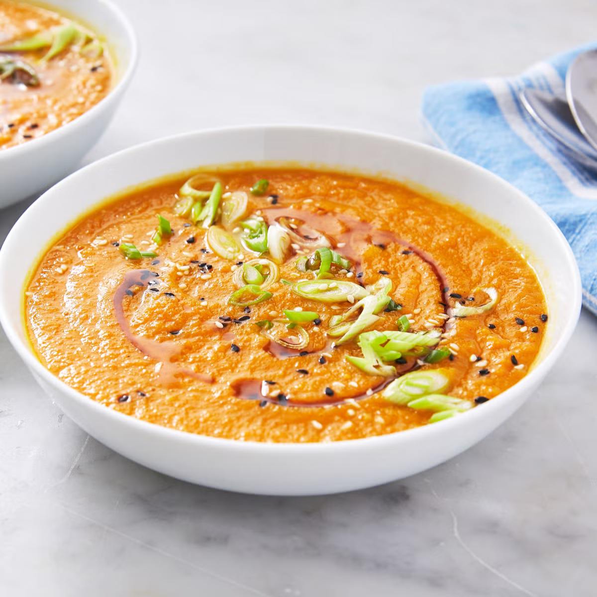 A bowl of vibrant orange carrot soup garnished with sesame seeds, chopped green onions, and a drizzle of soy sauce.