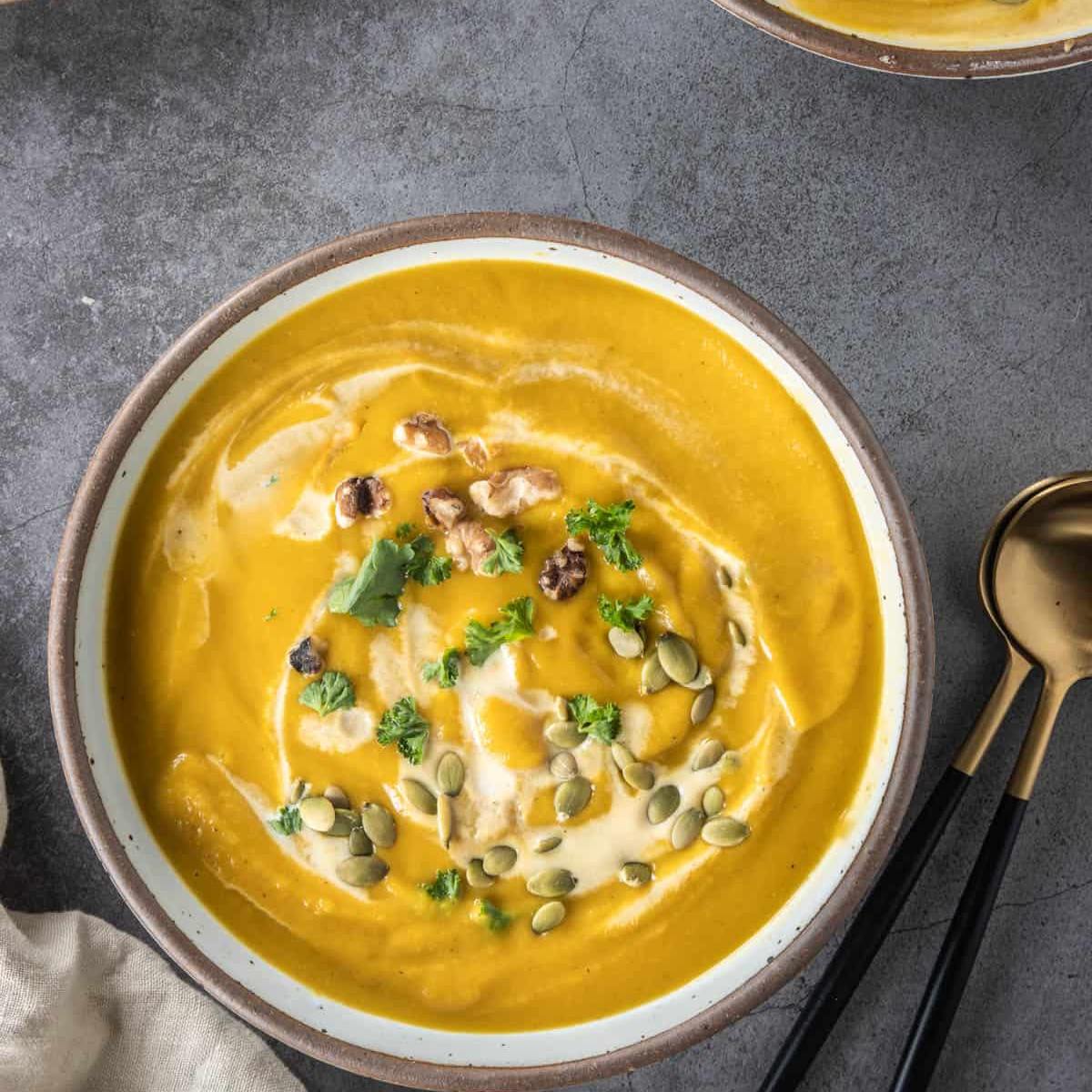 A bowl of creamy, golden pumpkin soup garnished with chopped walnuts, pumpkin seeds, parsley, and a swirl of cream.








