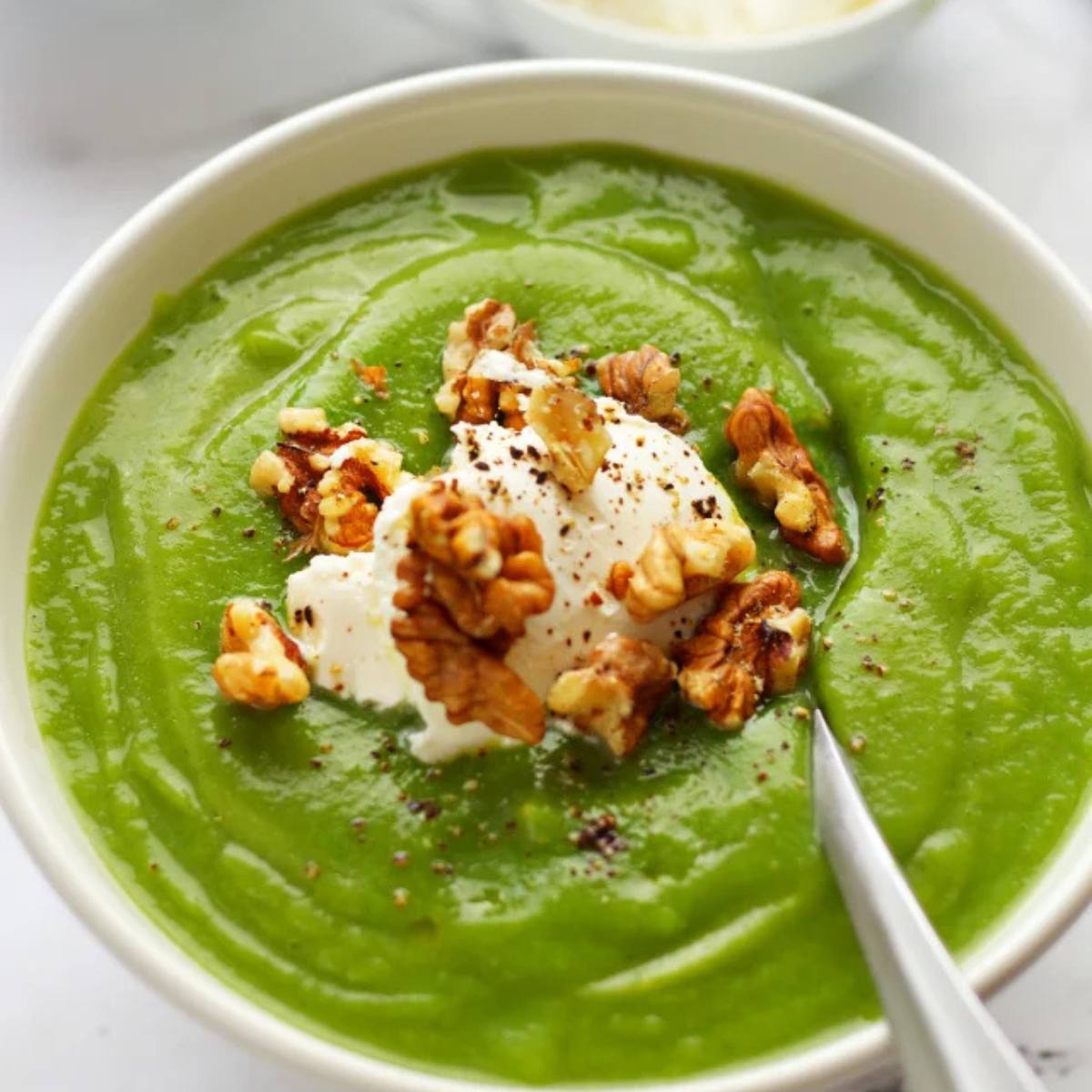 A bowl of vibrant green cream of spinach soup topped with dollops of sour cream, chopped walnuts, and a sprinkle of black pepper.