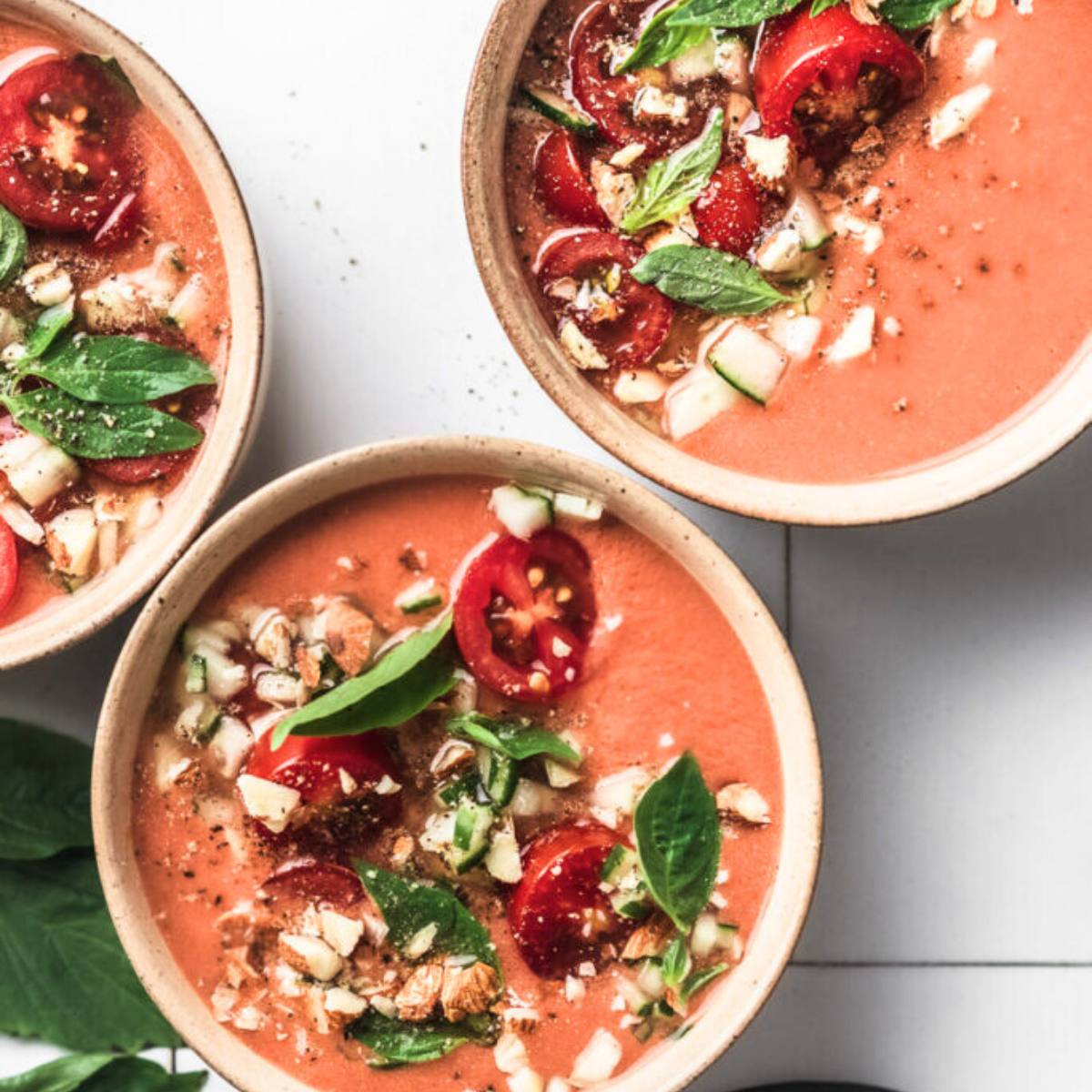 Three bowls of chilled gazpacho soup. The soup is a vibrant red color and is topped with chopped tomatoes, cucumber, almonds, and fresh basil leaves.