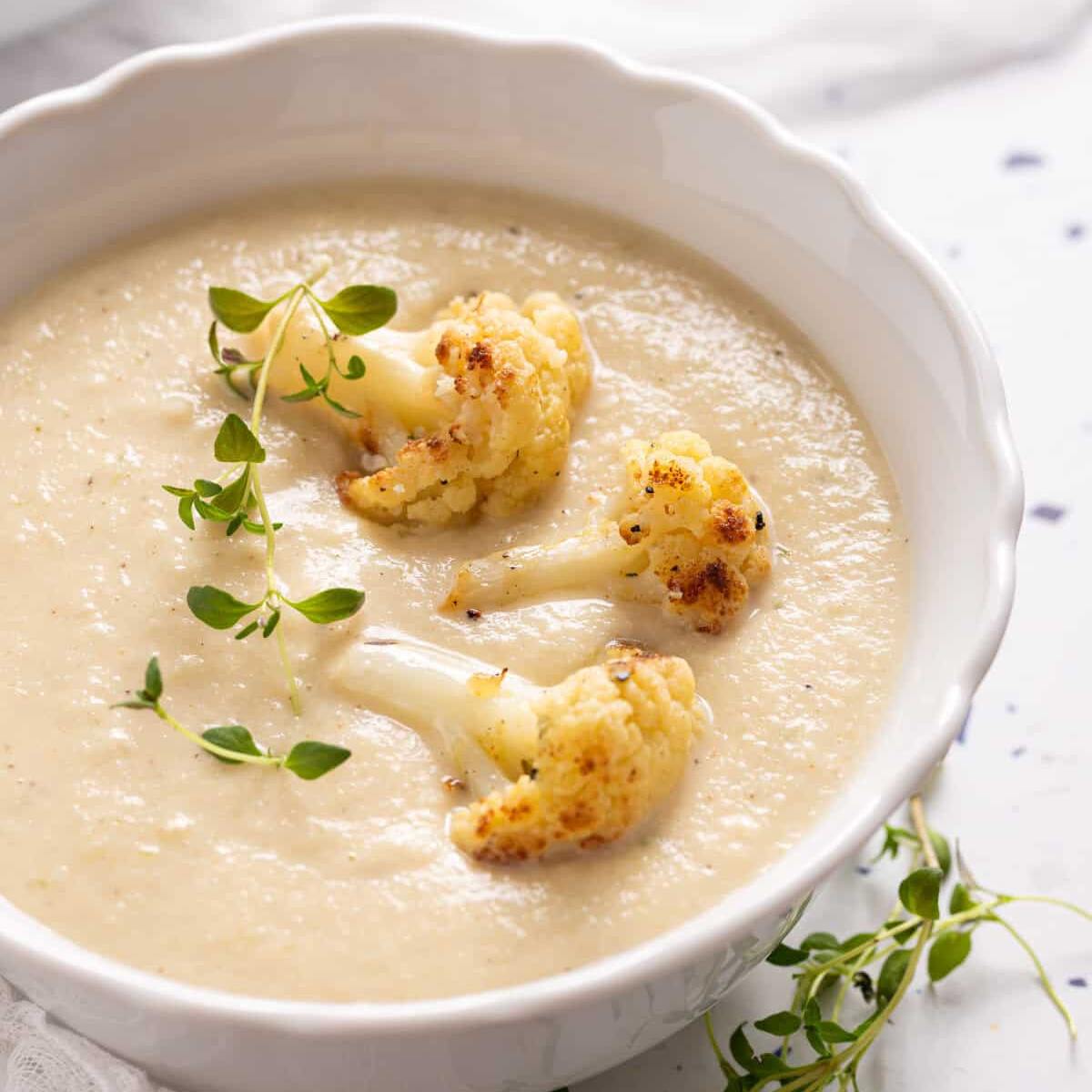 A bowl of creamy, pale yellow cauliflower soup with roasted cauliflower florets on top. The soup is garnished with fresh thyme sprigs.