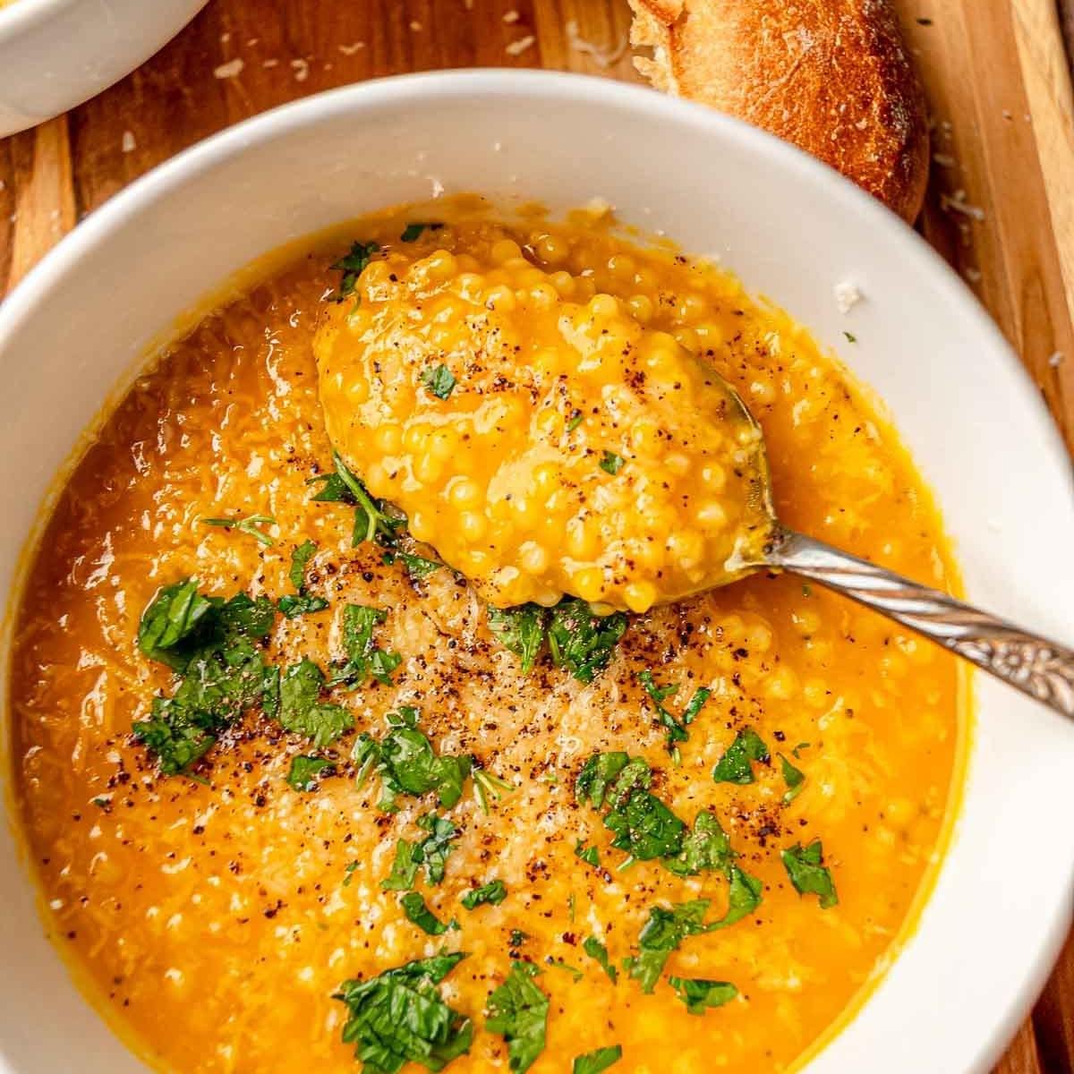 A bowl of creamy, golden soup with small, pearl-shaped pasta. The soup is garnished with fresh parsley and grated cheese. A spoon is scooping out the soup, and a crusty roll is visible in the background.