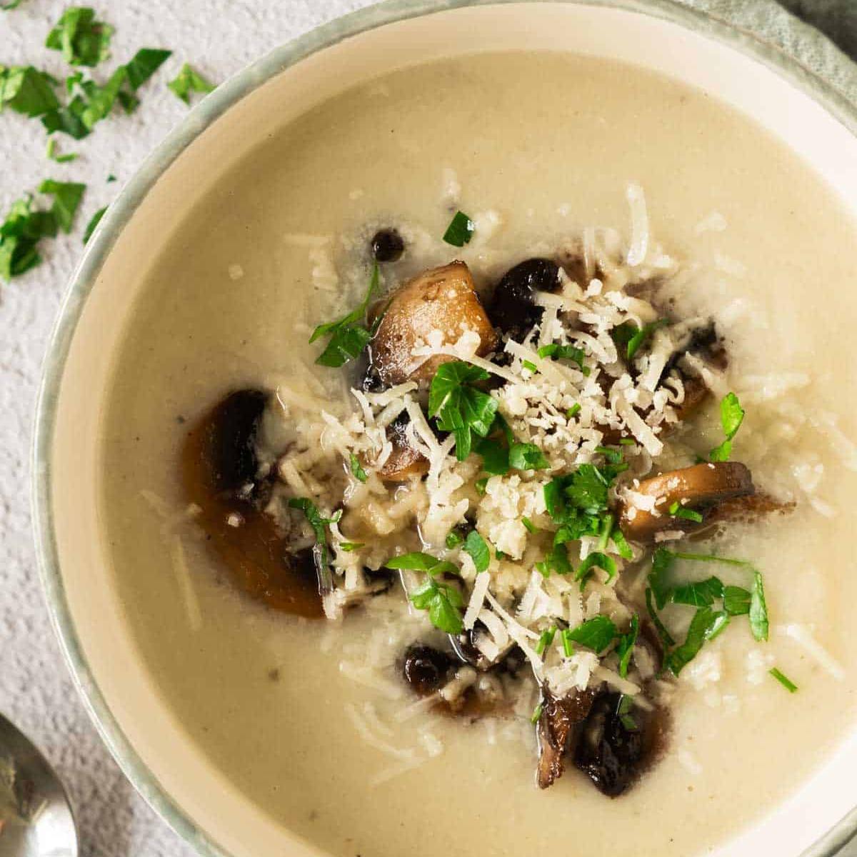 A bowl of creamy, pale yellow soup topped with sliced mushrooms, grated cheese, and chopped parsley.