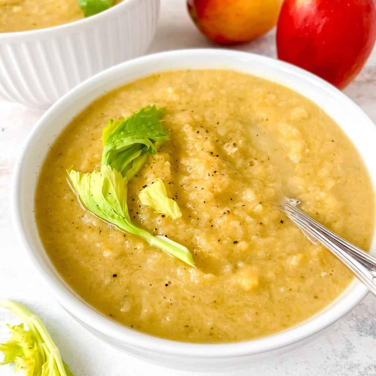 A bowl of creamy, golden soup garnished with fresh celery leaves. A spoon is resting in the soup, and a few apples are visible in the background.