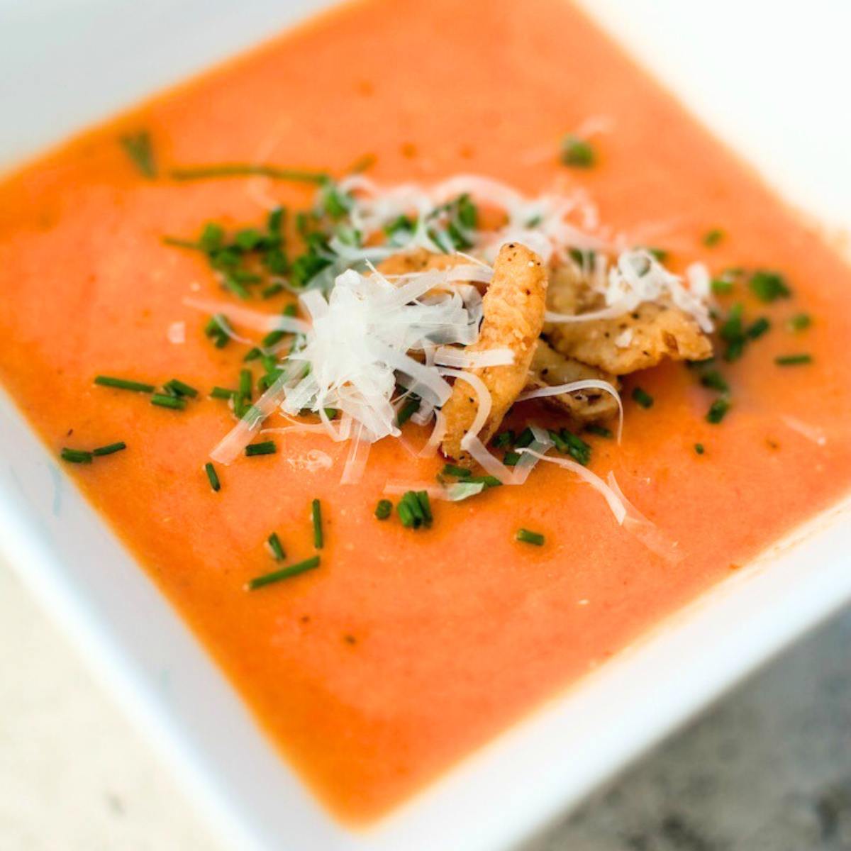 A bowl of creamy, vibrant orange tomato soup garnished with croutons, shredded cheese, and chives.