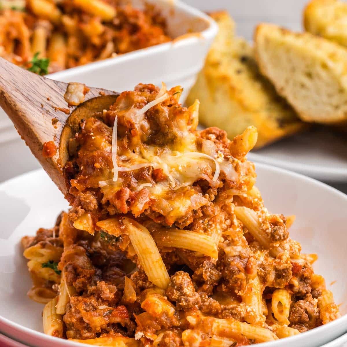 A close-up of cheesy beef pasta casserole with penne, ground beef, and marinara sauce, topped with melted cheese. A wooden spoon lifts a portion from the dish. 
