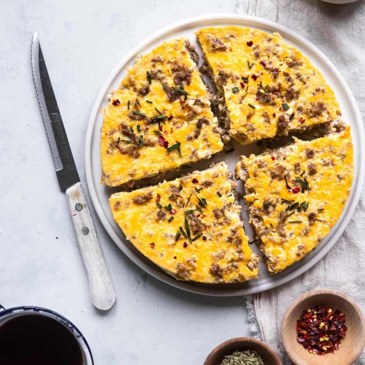 A sliced Instant Pot breakfast casserole with sausage and melted cheese on a white plate, garnished with herbs and chili flakes, served alongside spices and coffee.
