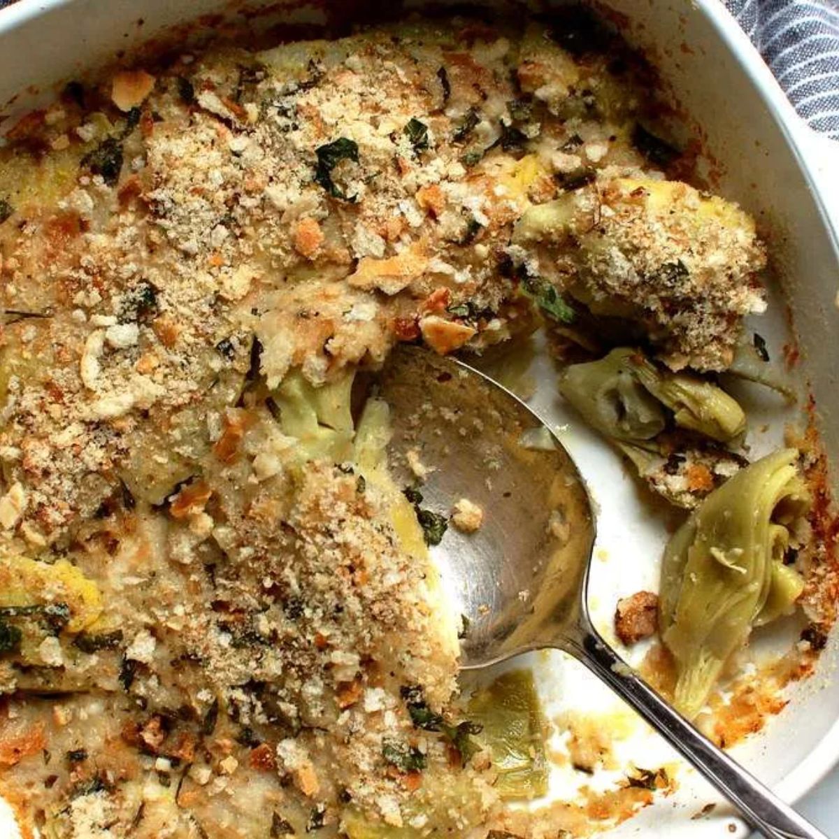 A baked artichoke casserole with a golden breadcrumb topping, garnished with herbs. A serving spoon reveals tender artichokes coated in a creamy, cheesy sauce.
