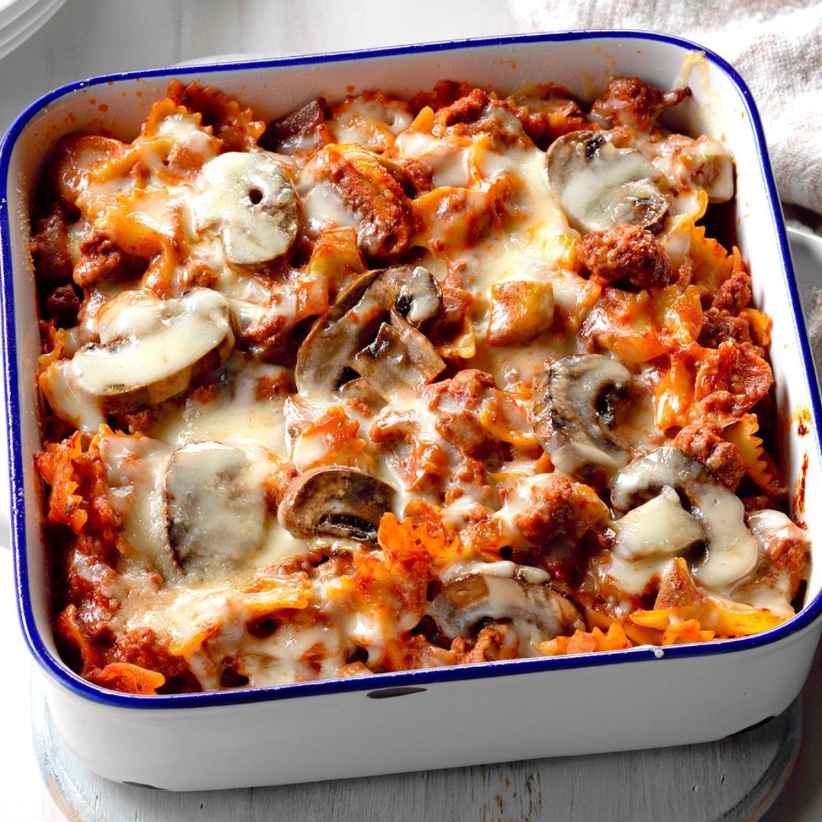 A baked pasta casserole with farfalle, ground beef, mushrooms, and melted mozzarella in a rich tomato sauce, served in a white enamel dish with blue trim.

