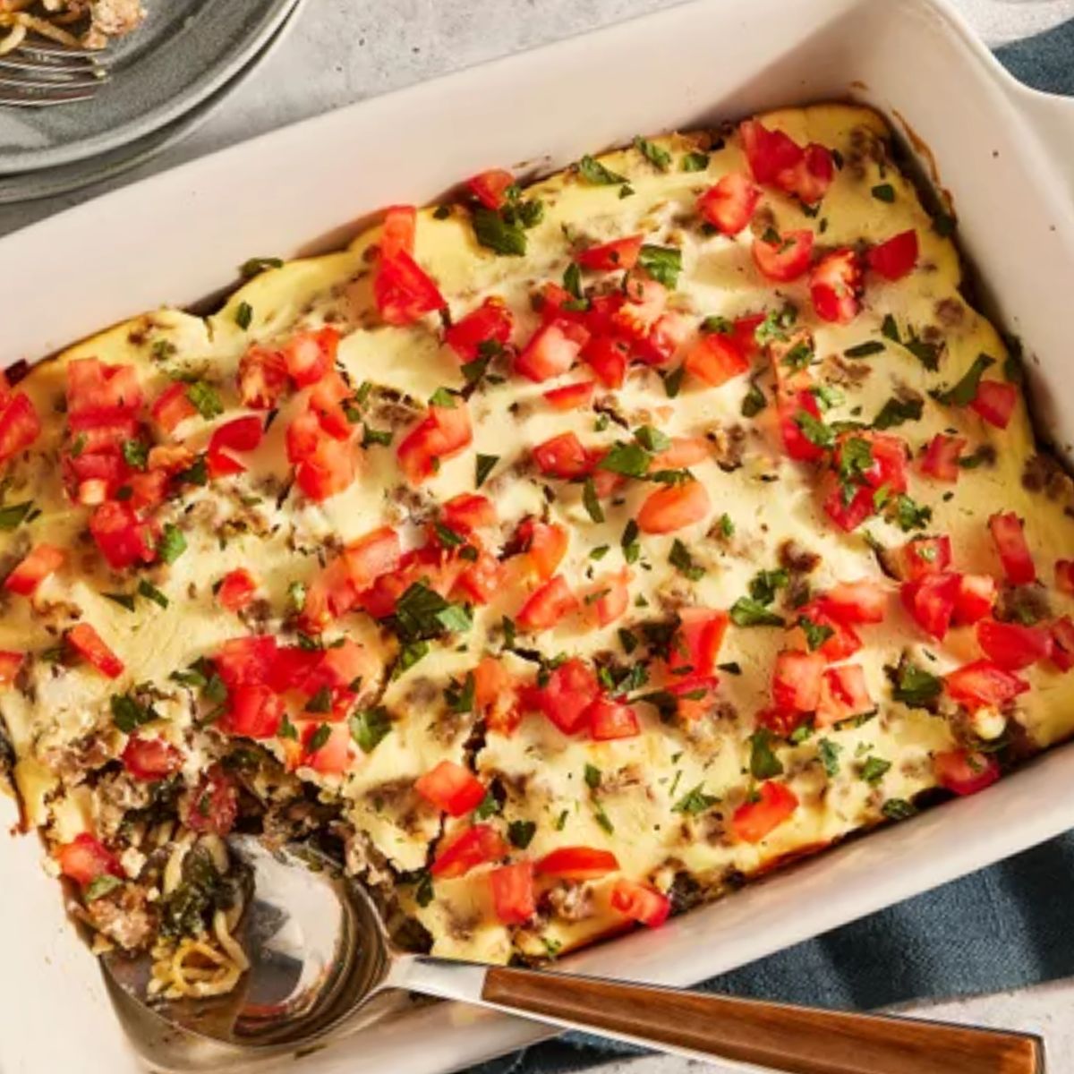 A baked Italian casserole with a creamy ricotta topping, spinach, mushrooms, and ground beef, garnished with diced tomatoes and parsley in a white baking dish.
