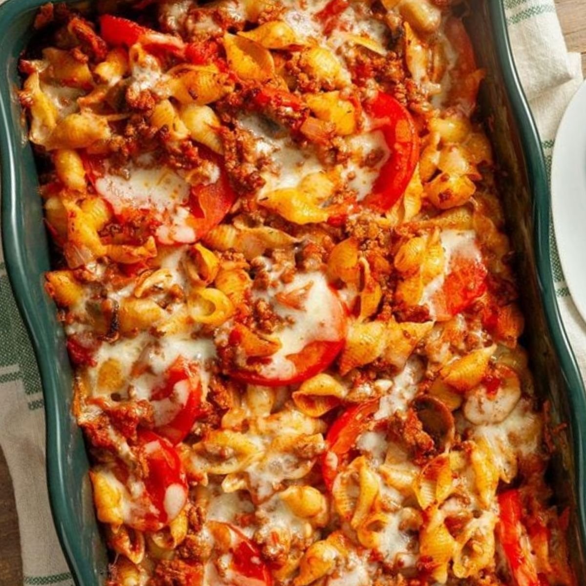 A baked pasta dish with shell pasta, ground beef, tomato sauce, and melted cheese, topped with fresh tomato slices and herbs in a green casserole dish.