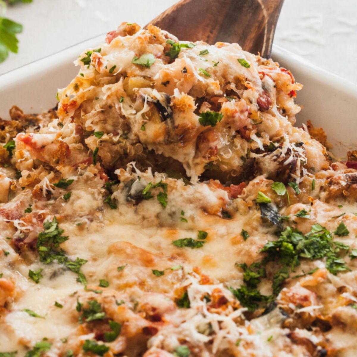 
A cheesy Italian quinoa casserole with roasted vegetables, tomato sauce, and fresh herbs, baked until golden brown, with melted cheese and chopped parsley on top. Served on a white baking dish. 
