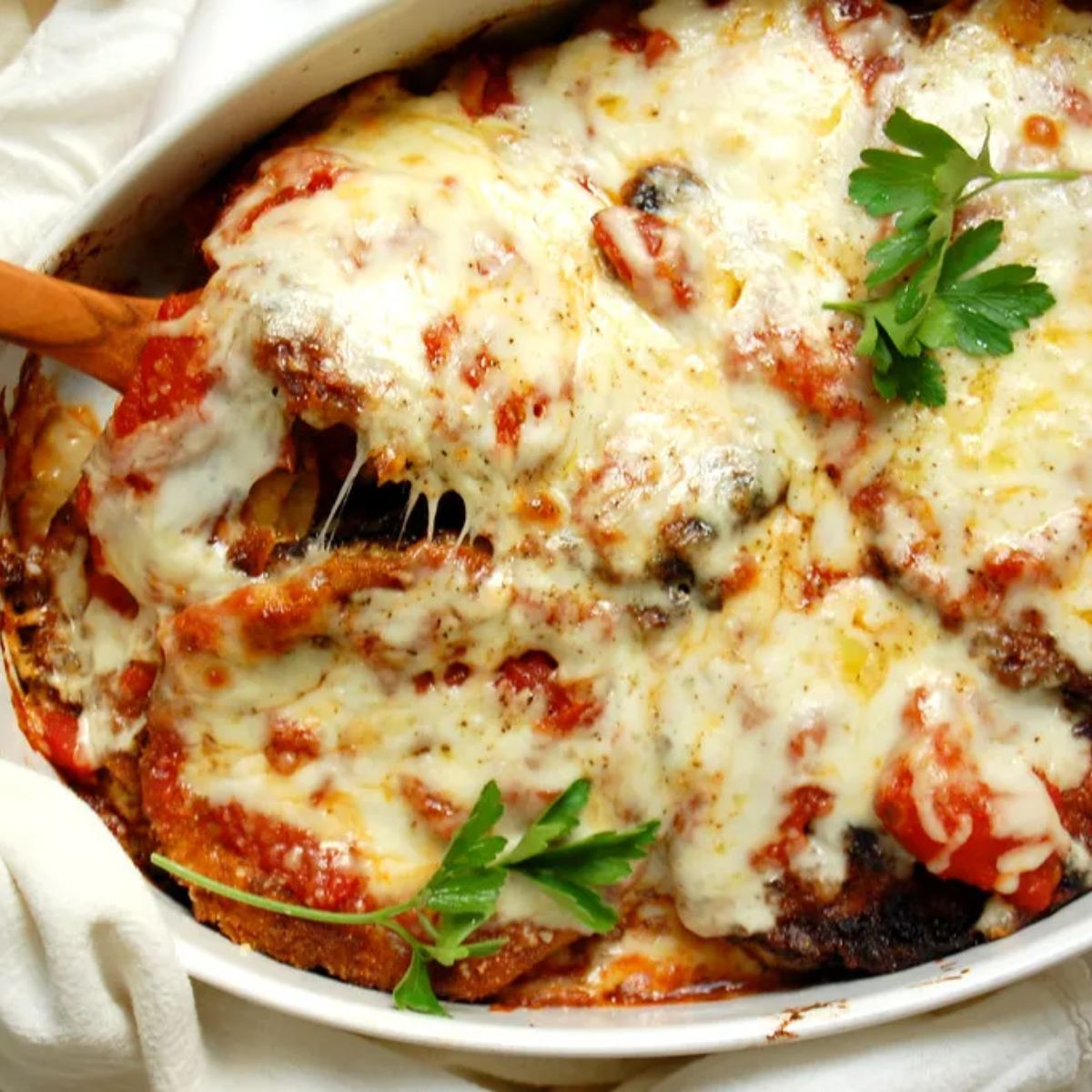 Baked eggplant Parmesan casserole in a white dish, covered in melted cheese and marinara sauce, garnished with fresh parsley. A wooden spoon lifts a portion.
