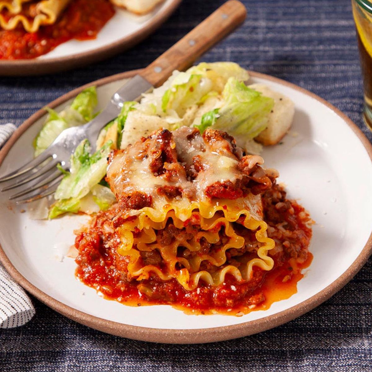 A plate of lasagna roll-ups with ruffled pasta edges, stuffed with meat and cheese, covered in marinara sauce, and topped with melted cheese. Served with salad and bread.