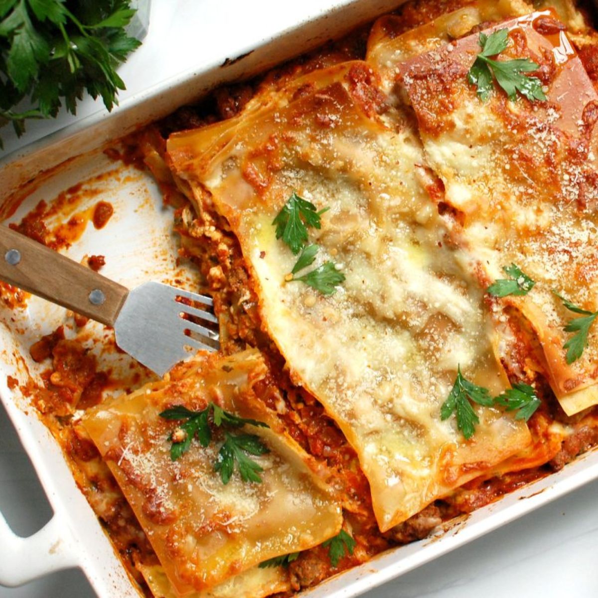 A freshly baked lasagna in a white casserole dish with layers of pasta, meat sauce, and melted cheese in a golden, bubbly crust topped with parsley.