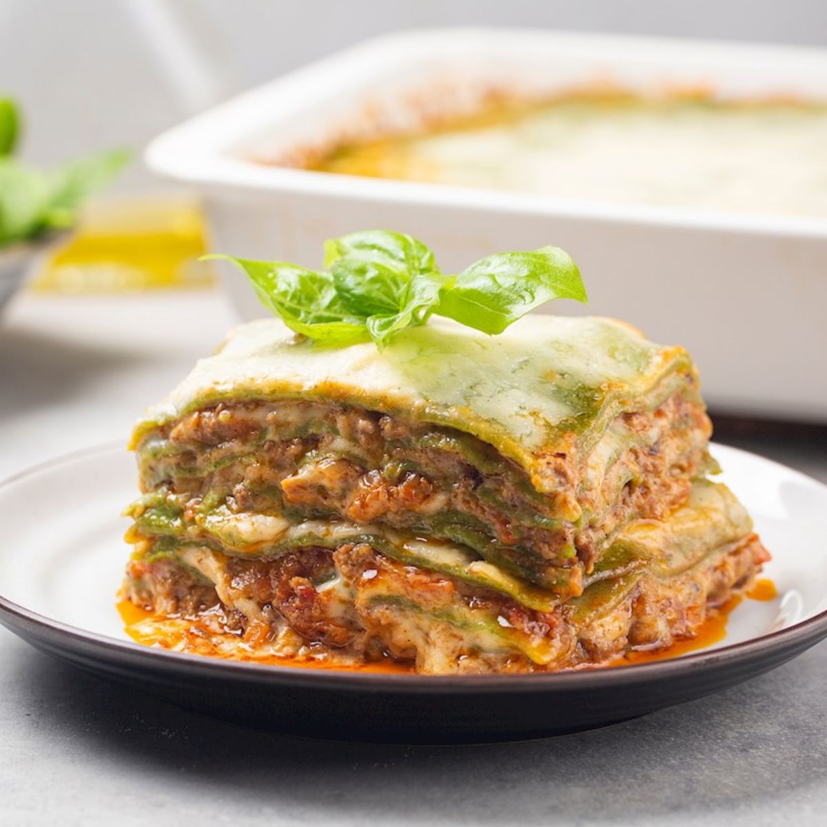 A slice of spinach pasta lasagna layered with rich meat sauce, creamy béchamel, and melted cheese, garnished with fresh basil, served on a white plate.
