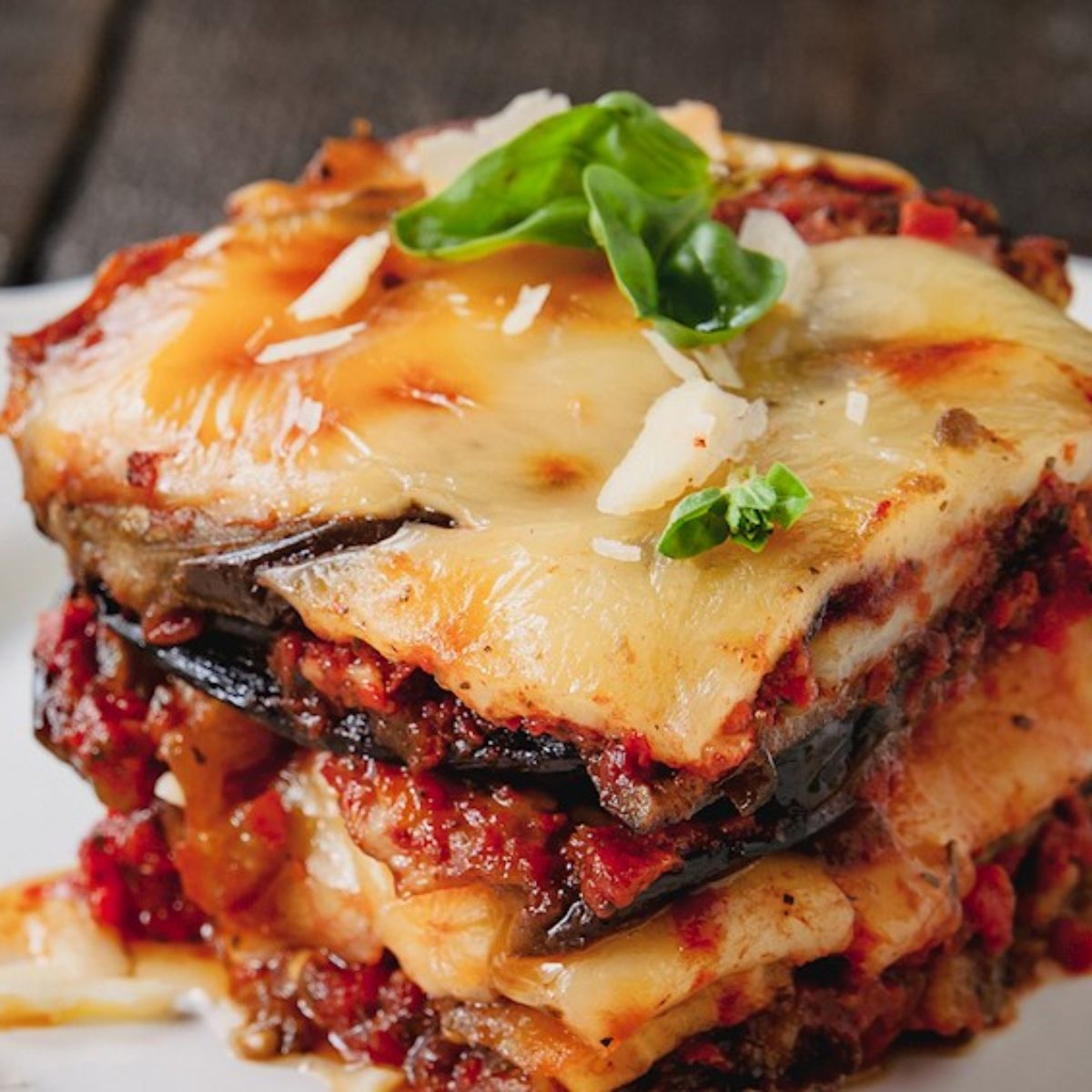 A slice of Parmigiana alla Napoletana with layers of fried eggplant, rich tomato sauce, melted cheese, and fresh basil, served on a white plate.
