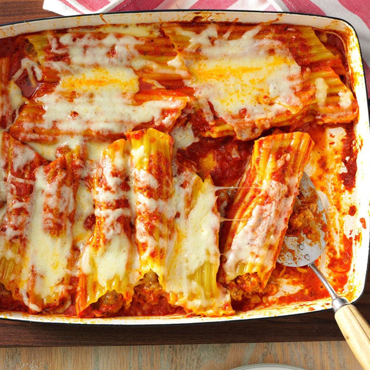 A baked dish of sausage-stuffed manicotti covered in marinara sauce and melted cheese, served in a white casserole dish with a portion scooped out.
