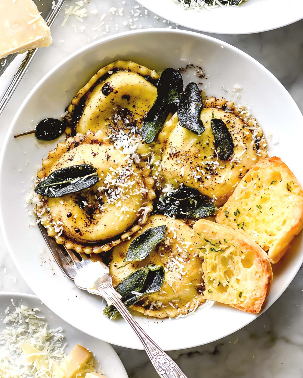 A white bowl of browned butter sage ravioli served with garlic bread, topped with crispy sage leaves and grated cheese for an easy fall dinner.