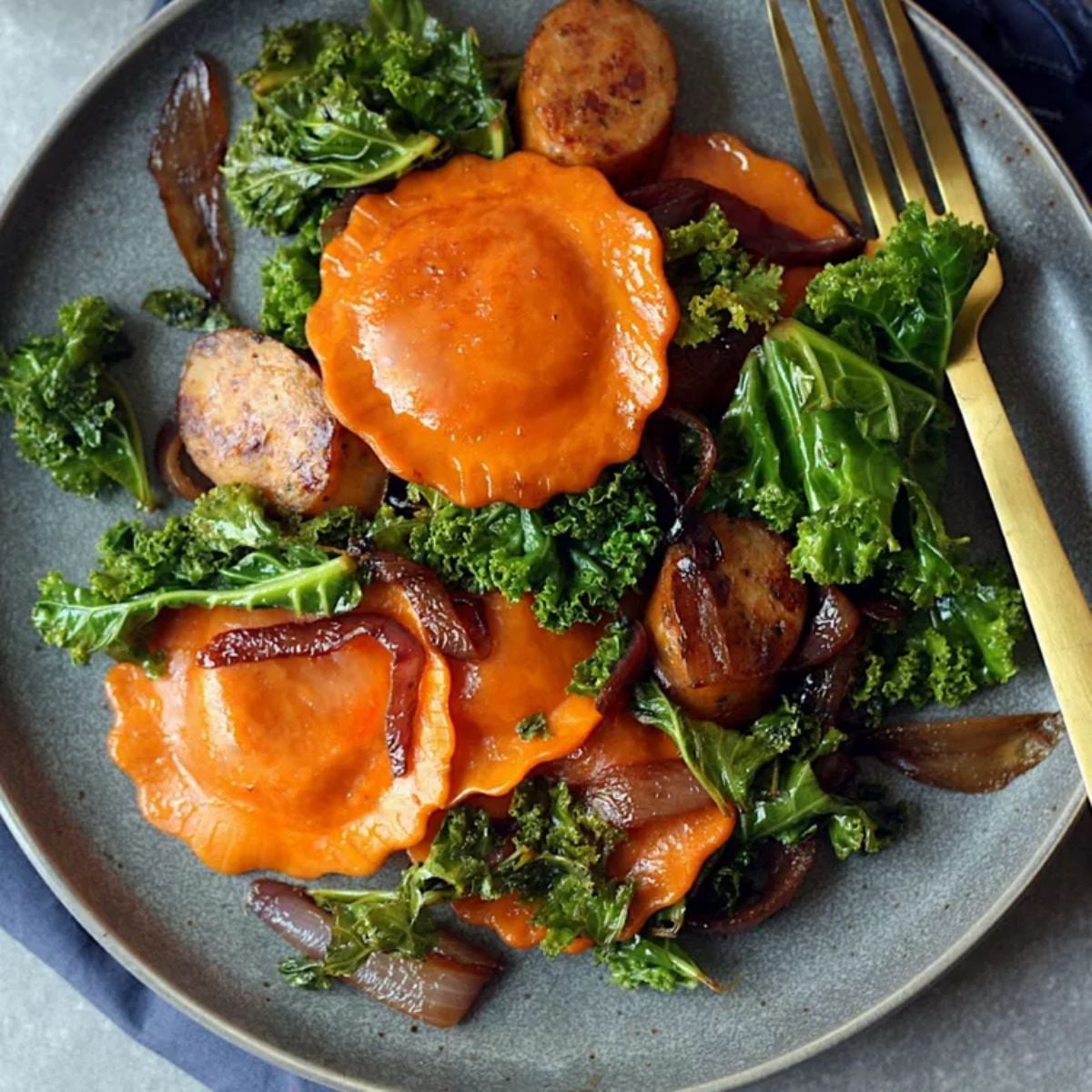 Stuffed pasta rounds mixed with caramelized onions, sausage slices, and tender greens on a gray plate.
