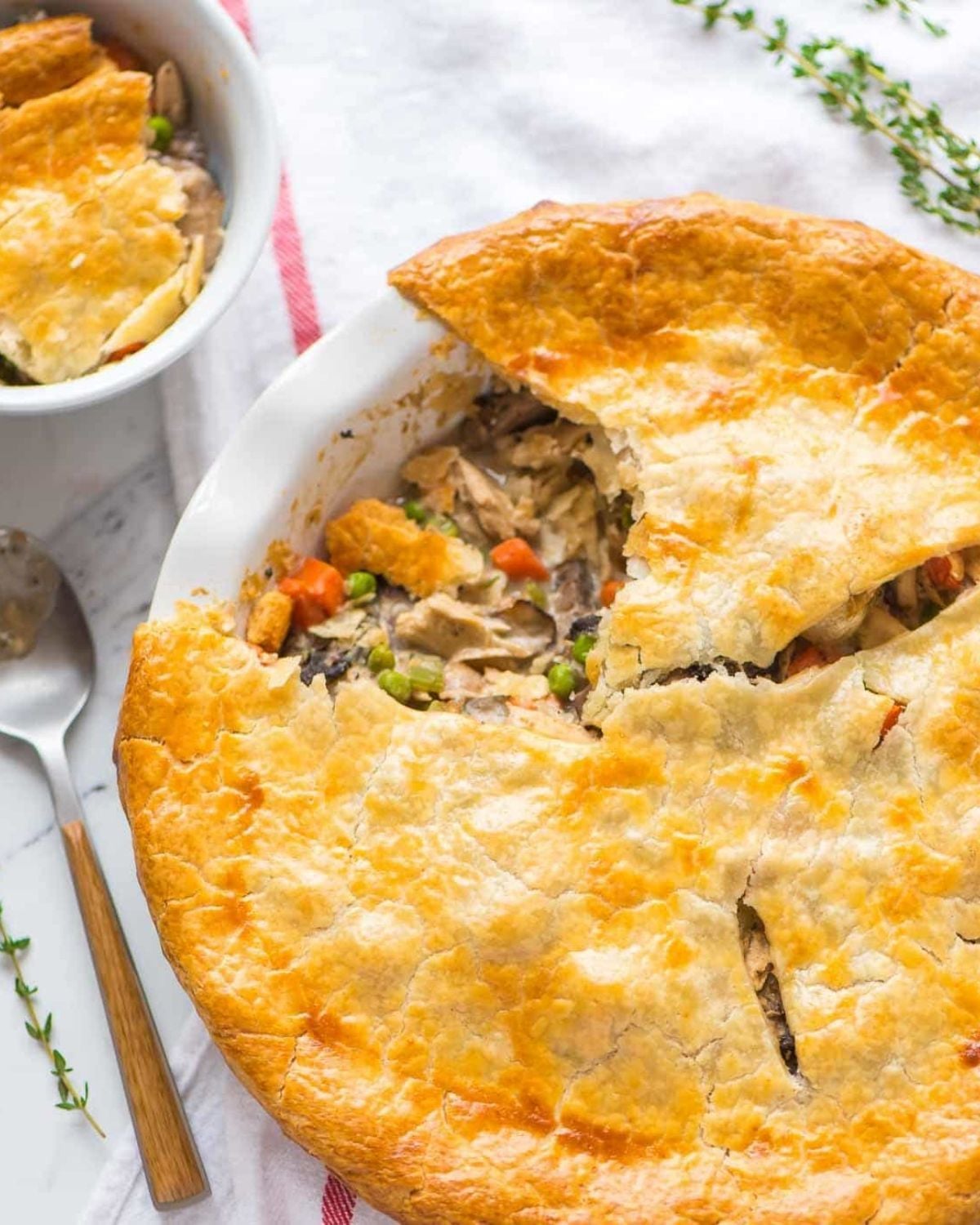 A freshly baked chicken pot pie in a white dish with a slice cut open to reveal a creamy filling of chicken, carrots, and peas, alongside a smaller portion in a ramekin.