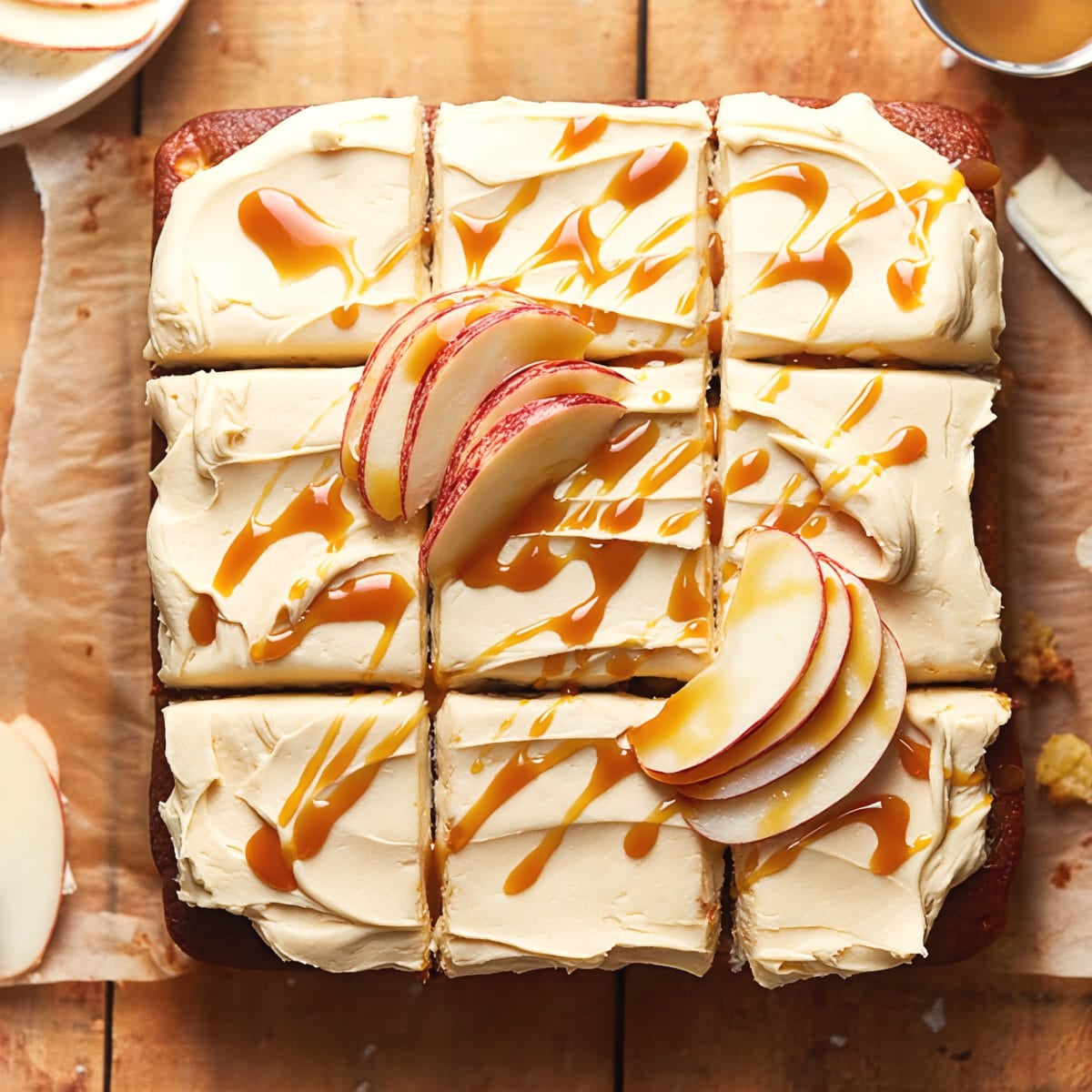 A top-down view of a square apple cake cut into nine individual slices, frosted with pale caramel-colored icing, drizzled with glossy amber caramel sauce, and garnished with fresh apple slices on parchment paper over a wooden table.
