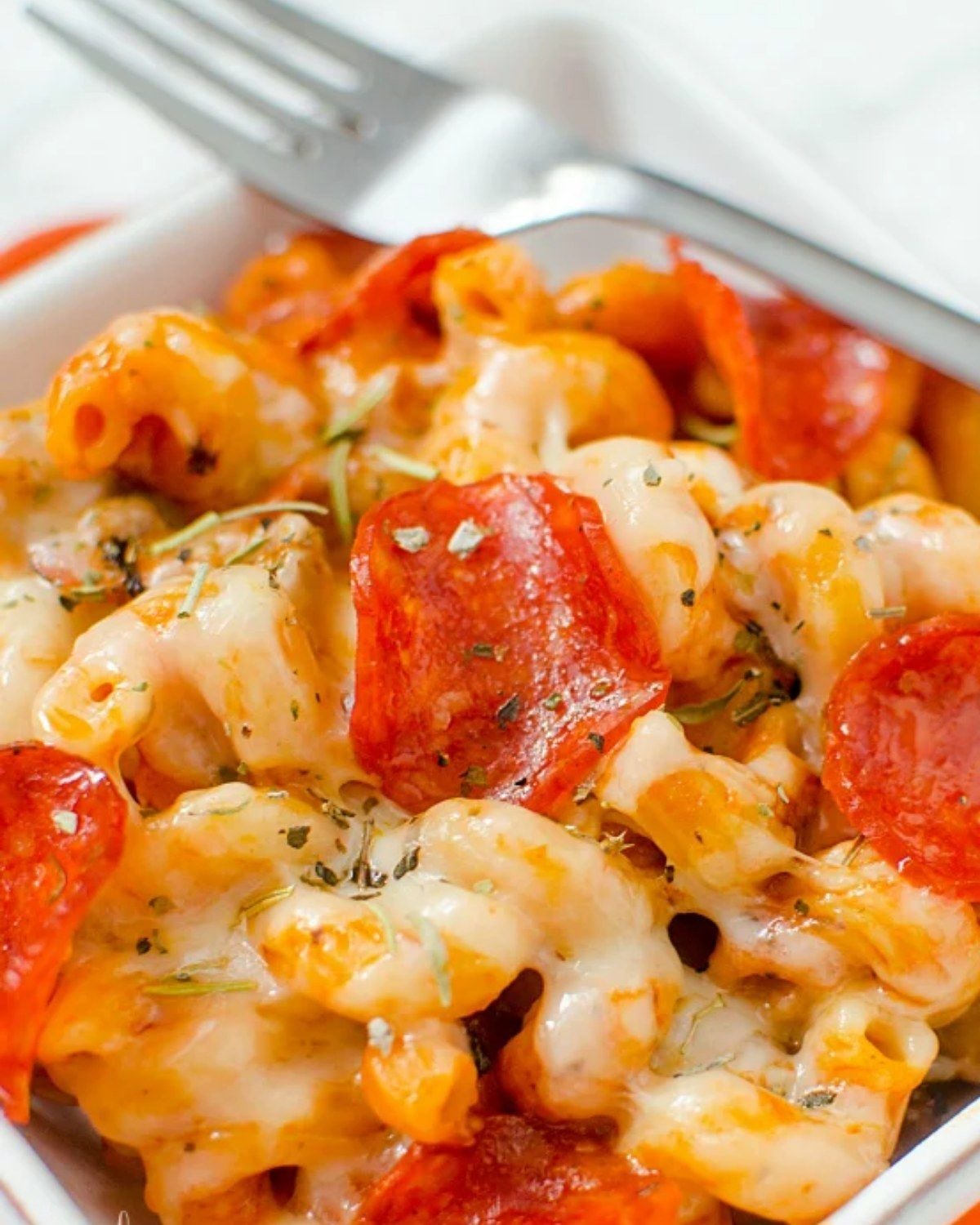 A white bowl of cheesy pizza pasta loaded with pepperoni slices and herbs, with a fork resting on the edge
