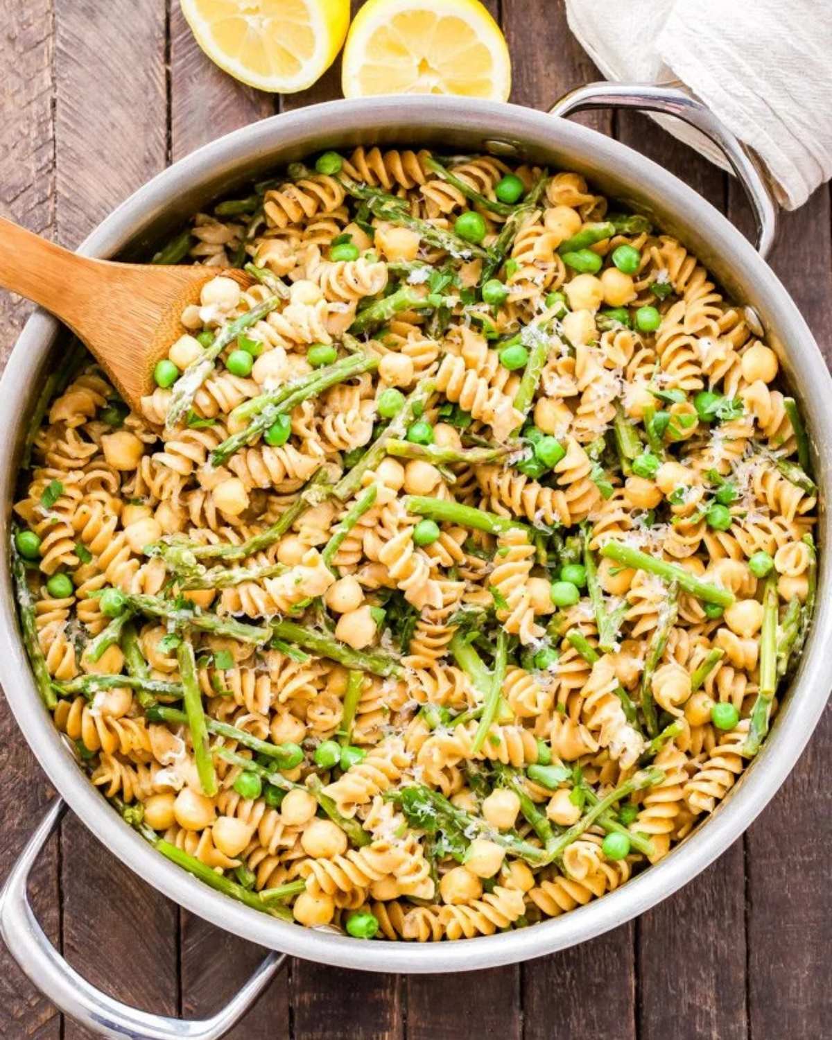A large skillet filled with rotini pasta, asparagus spears, green peas, and chickpeas tossed in a creamy sauce, with fresh lemons sitting nearby on a wooden surface.