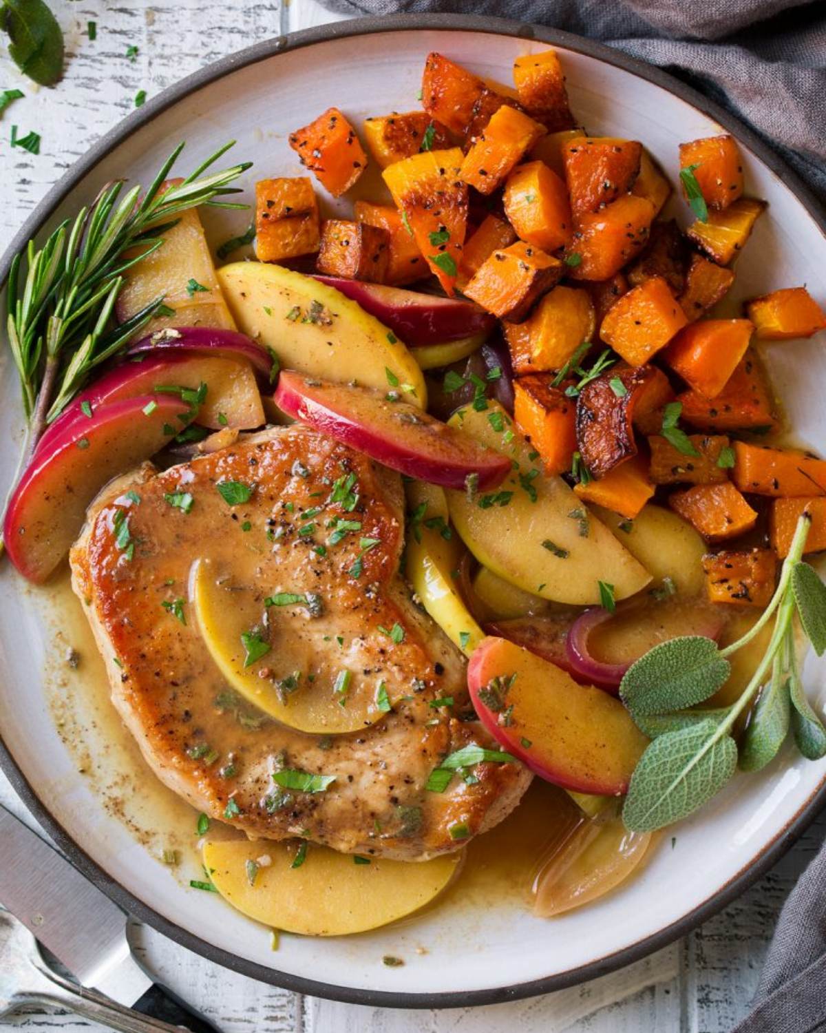 Seared meat chop served with glazed apple wedges, roasted squash cubes, and fresh herbs on a plate.