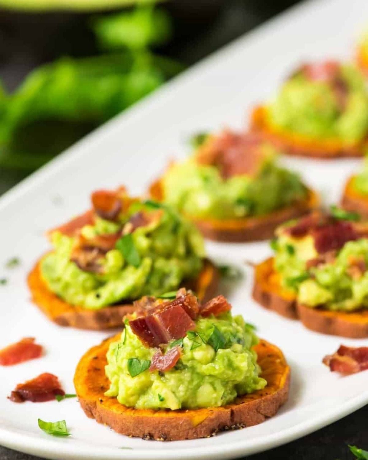 A close-up shot of roasted sweet potato rounds topped with mashed avocado and crispy bacon bits, arranged on a white serving platter.