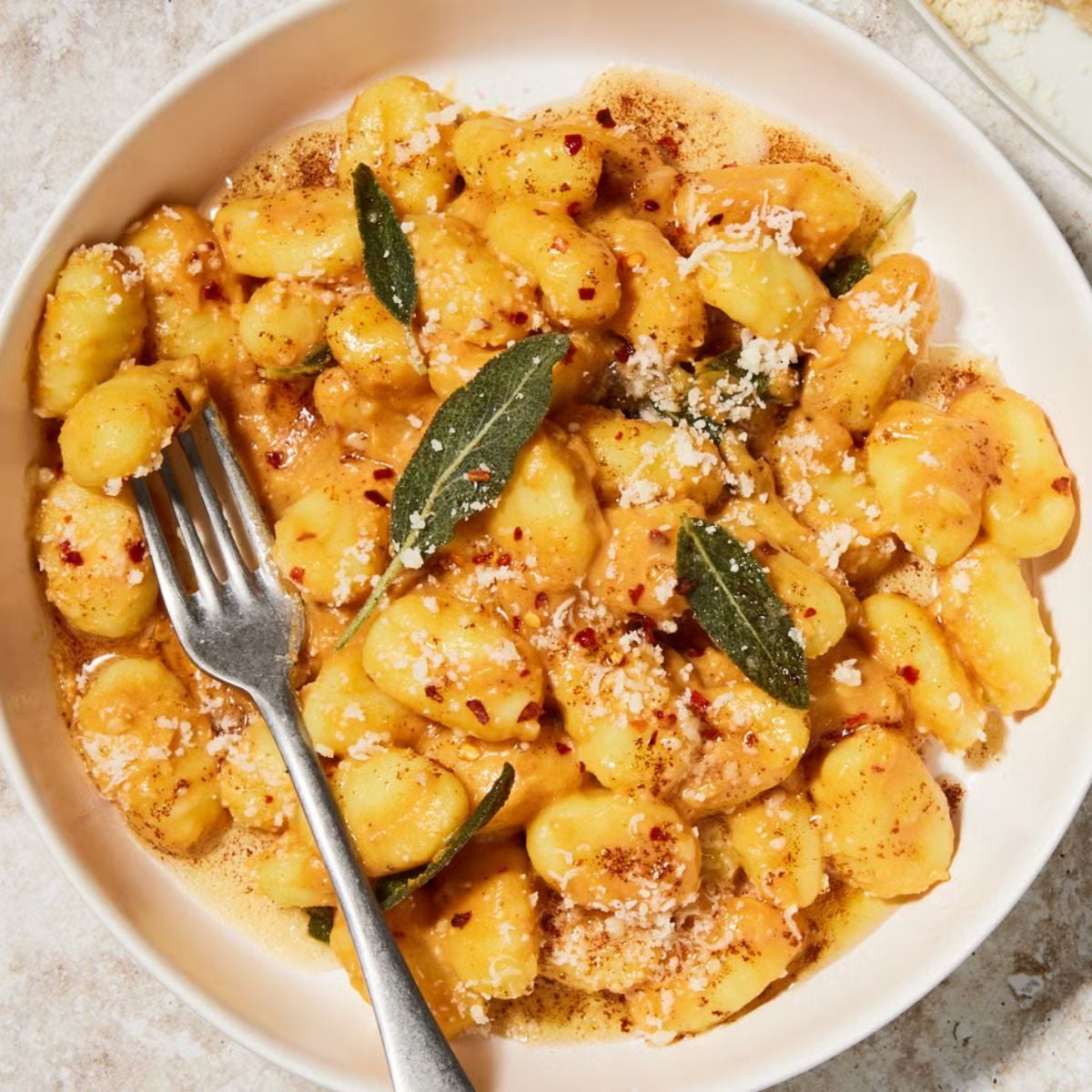 Golden dumpling pieces coated in a creamy sauce, garnished with grated cheese, red pepper flakes, and fresh sage leaves inside a bowl.