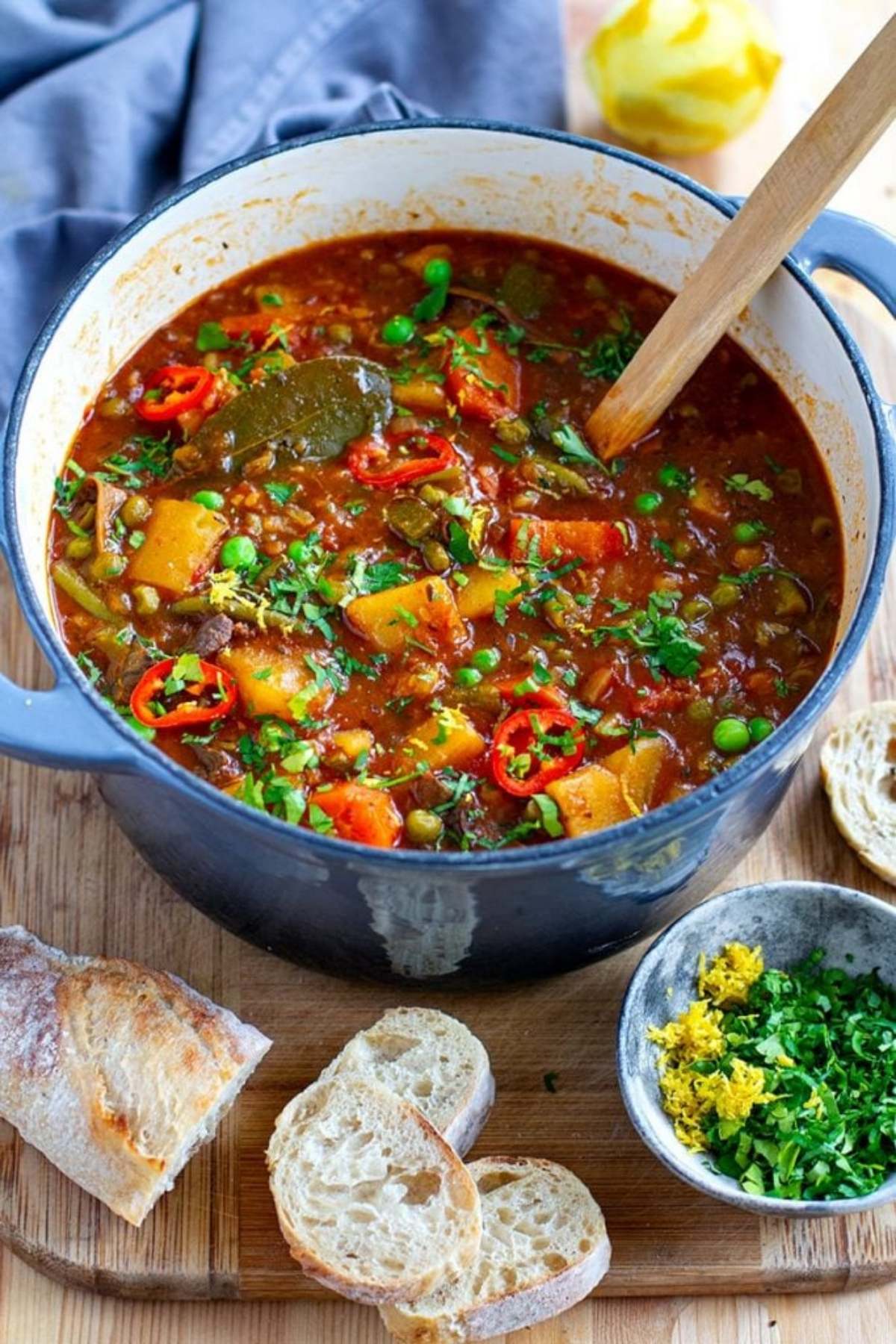 A blue pot filled with vegetable stew, garnished with fresh herbs and sliced red chilies, sitting on a wooden cutting board next to sliced bread and a bowl of herbs.