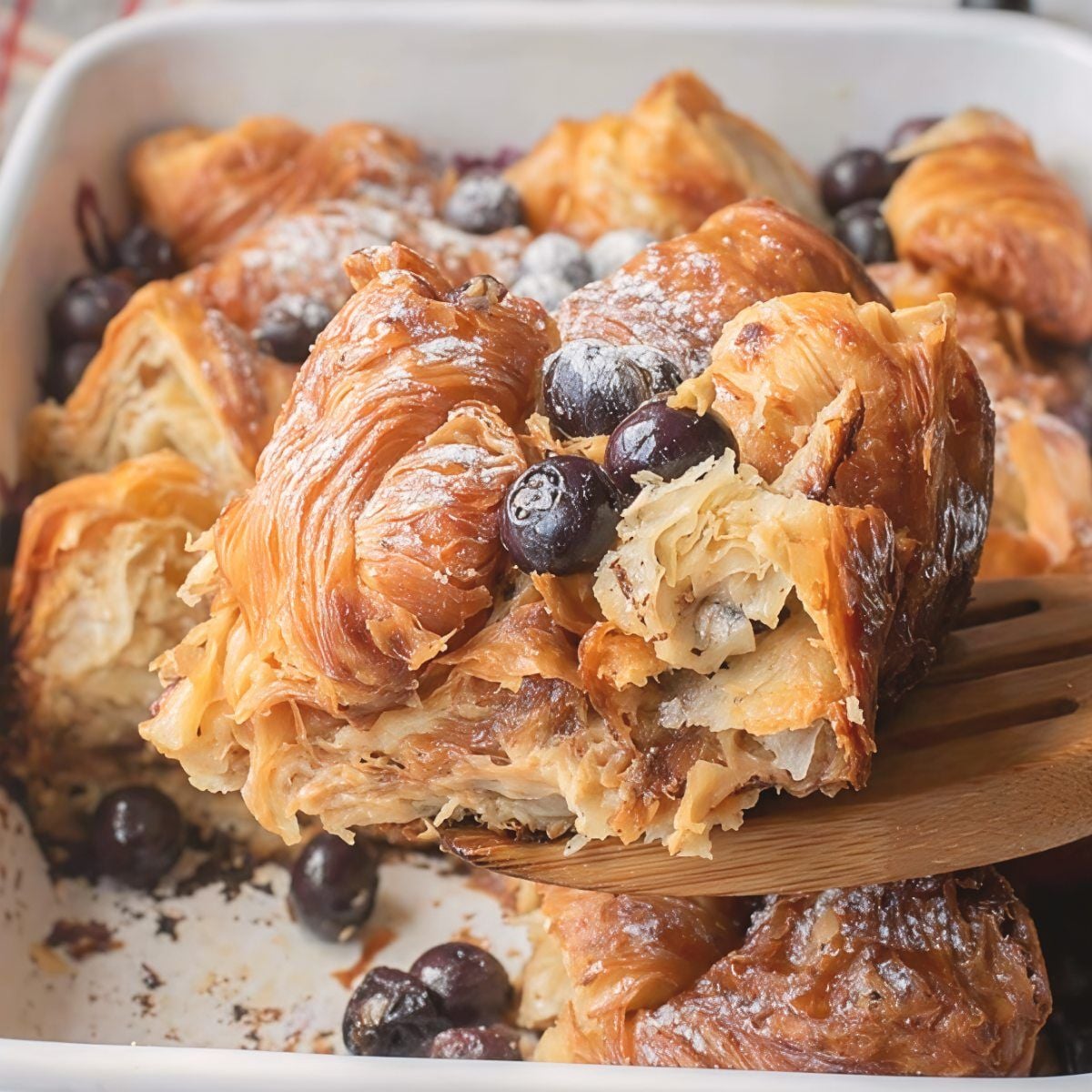 A wooden spatula lifting a slice of homemade blueberry croissant bake dusted with powdered sugar, a popular sweet brunch potluck idea.