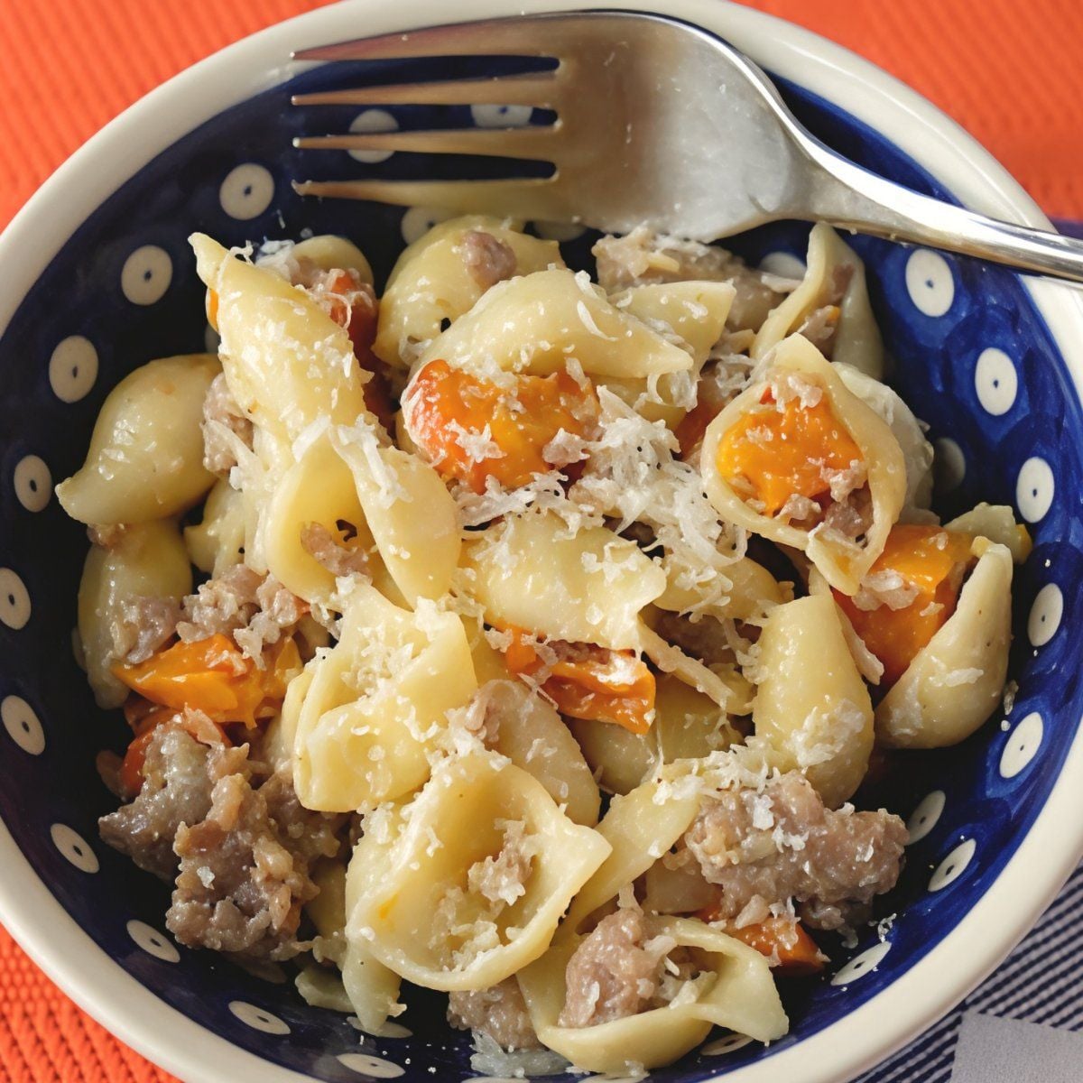 A blue and white polka dot bowl of burrata pasta with sausage and squash, topped with grated cheese and a fork, making an easy fall dinner.