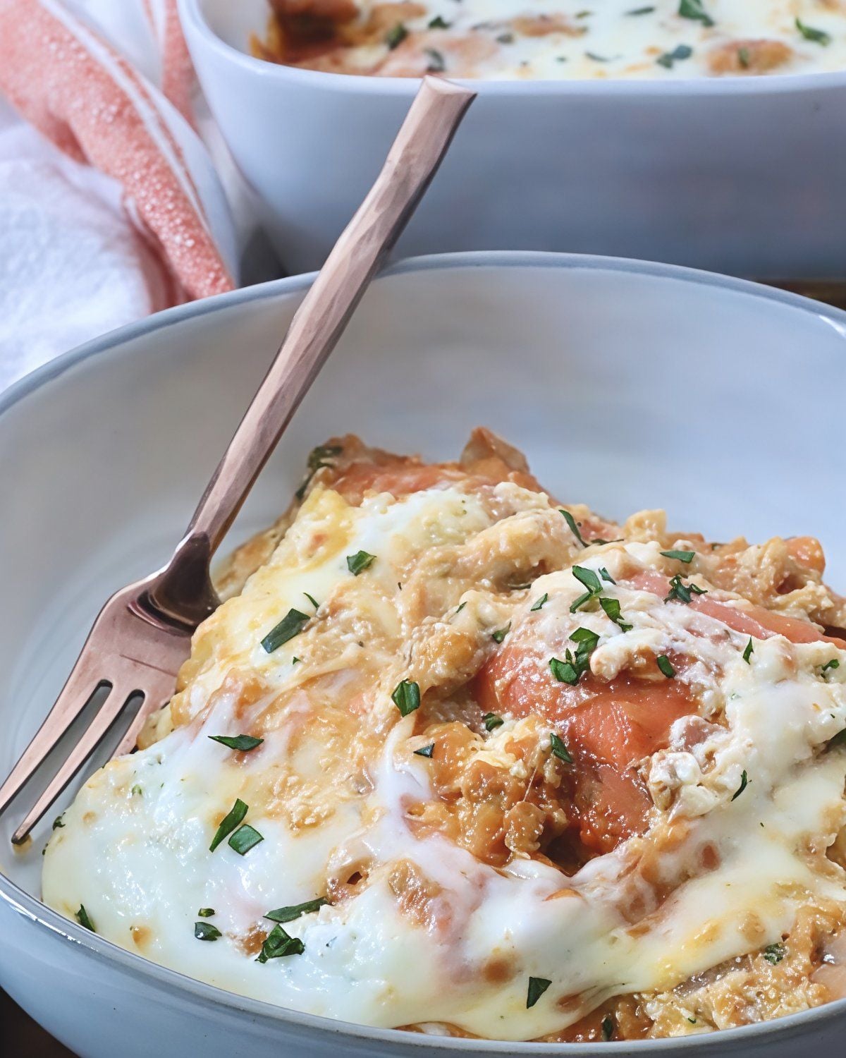 A serving of butternut squash baked ravioli in a white bowl with a fork, topped with melted cheese and fresh herbs for an easy fall dinner.