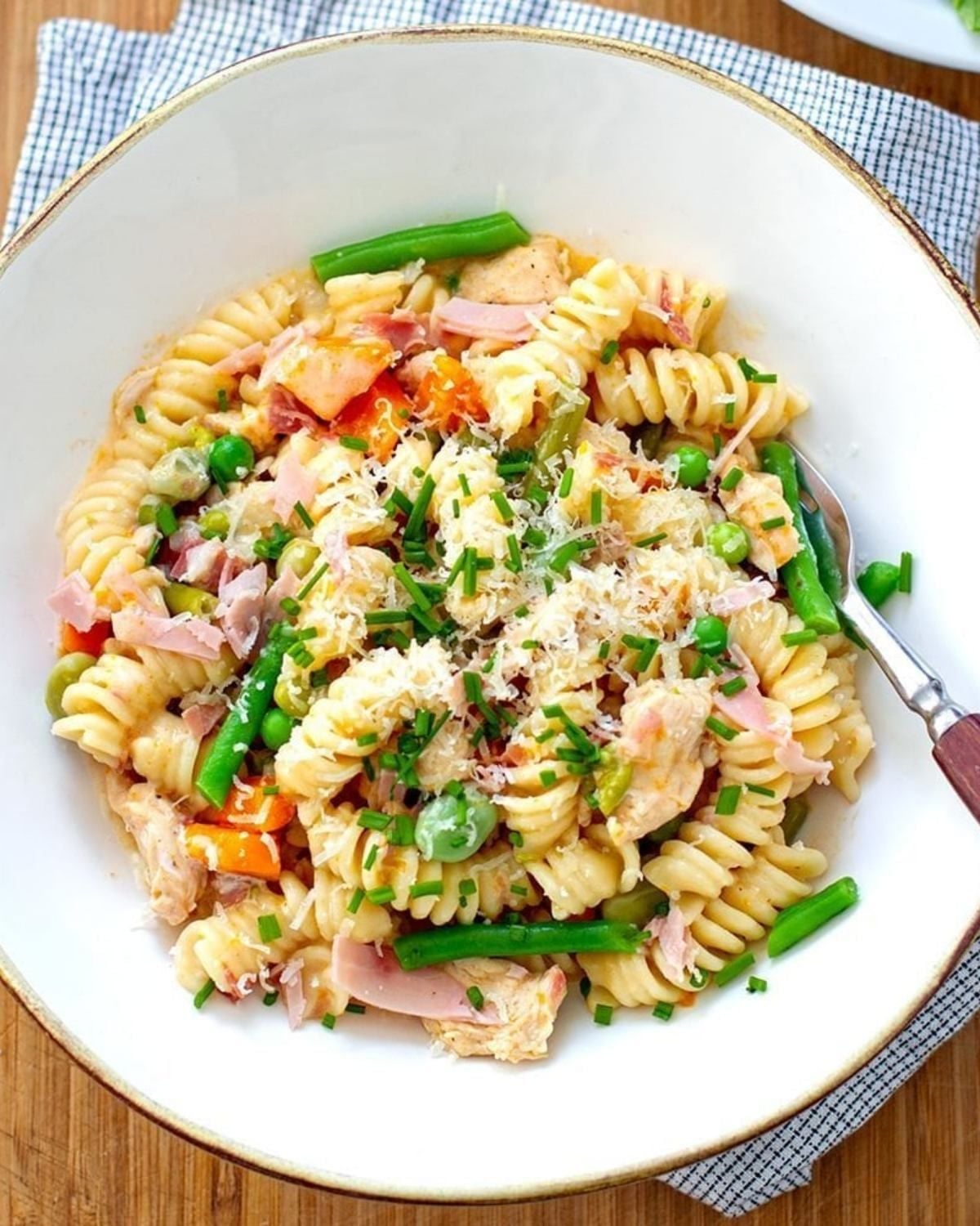 A close up view of a white bowl filled with chicken, ham, and vegetable pasta made with rotini, green beans, peas, and chicken pieces, garnished with grated cheese and chopped chives.