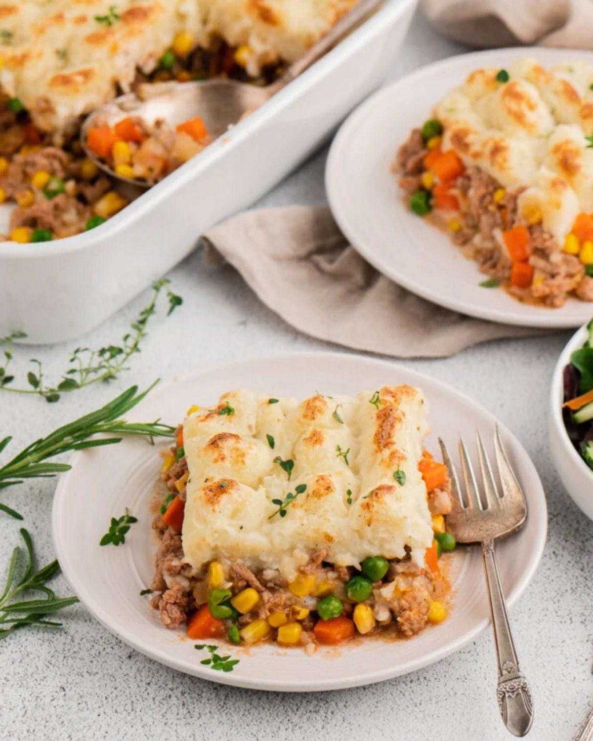 A slice of shepherd's pie with a golden mashed potato topping and a filling of meat, carrots, and peas on a white plate, with a baking dish of the pie in the background.