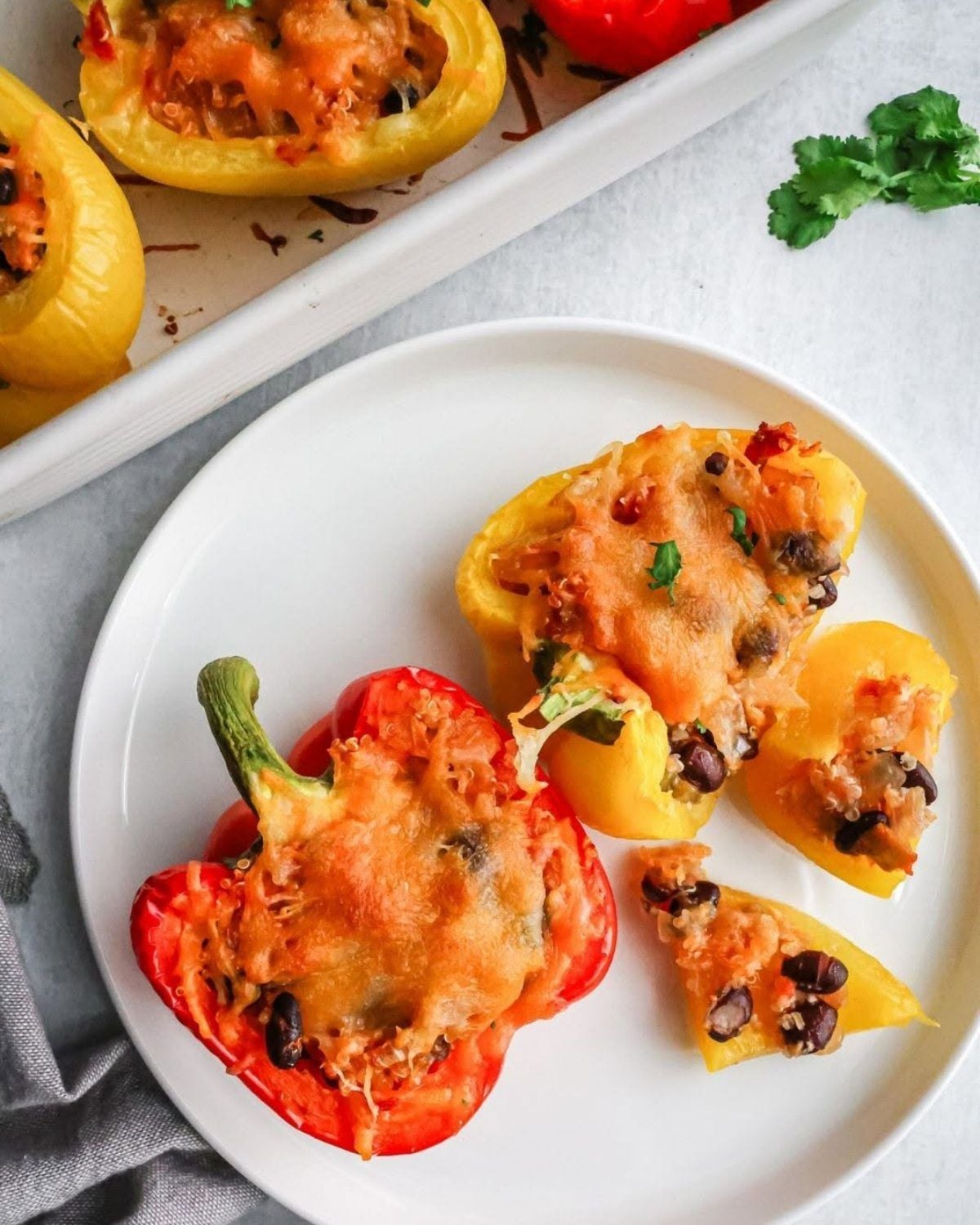 A top-down view of roasted red and yellow bell peppers stuffed with a savory mixture of black beans, grains, and melted cheese on a white plate, with a baking dish of more stuffed peppers in the background.